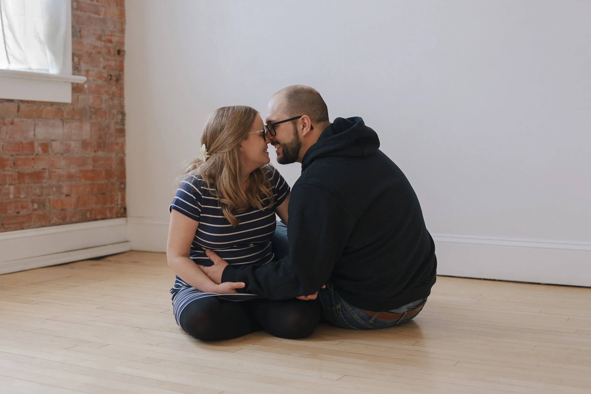 A young woman and a man sitting on a wooden floor with their foreheads touching, smiling, in a room with a brick wall and white wall, sharing an intimate moment.