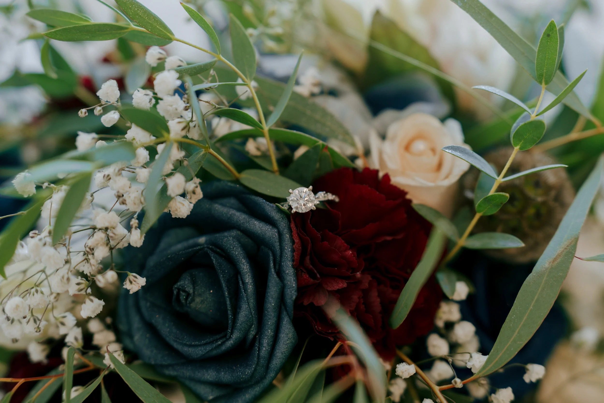 Close-up of a bouquet featuring navy blue and burgundy roses, white baby's breath, and greenery, with a diamond engagement ring placed on the navy rose.