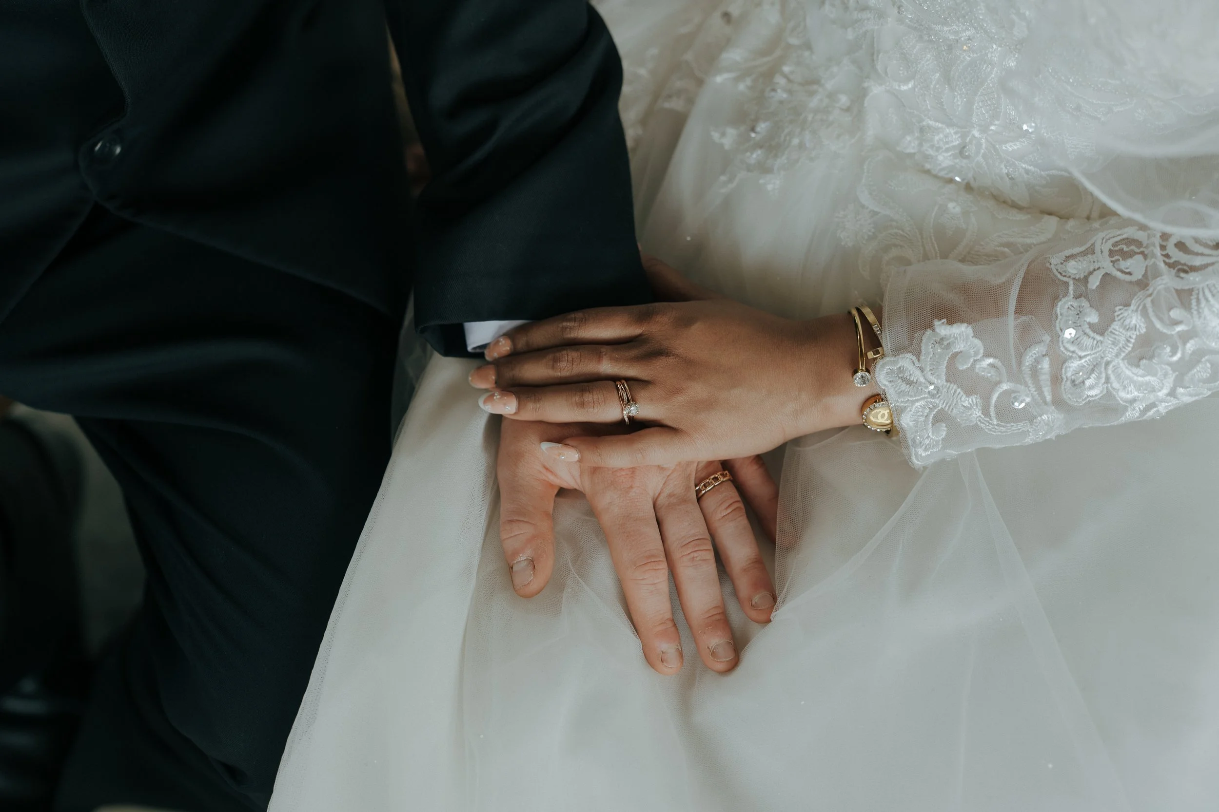 Close-up of a bride and groom holding hands during a wedding, showing wedding rings on their fingers. The bride's hand is resting on the groom's arm, and she is wearing a lace wedding dress and gold jewelry.