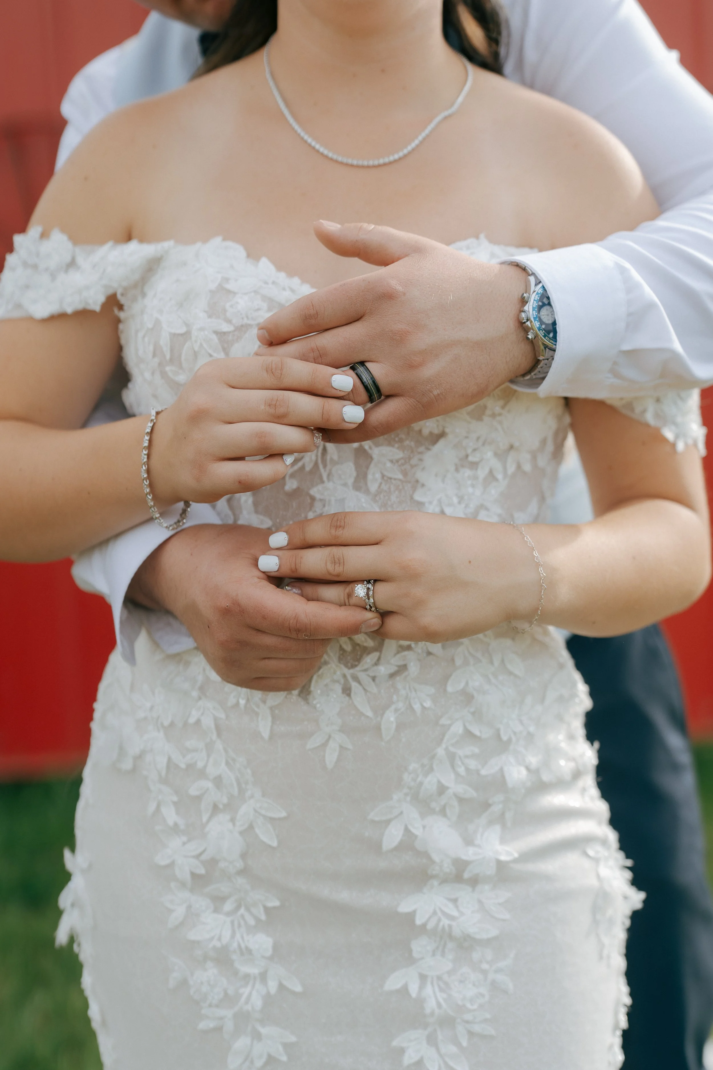 Close-up of a wedding couple's hands and torsos, with the bride wearing a lace wedding dress and jewelry, and the groom wearing a white shirt and watch, both showing wedding rings.