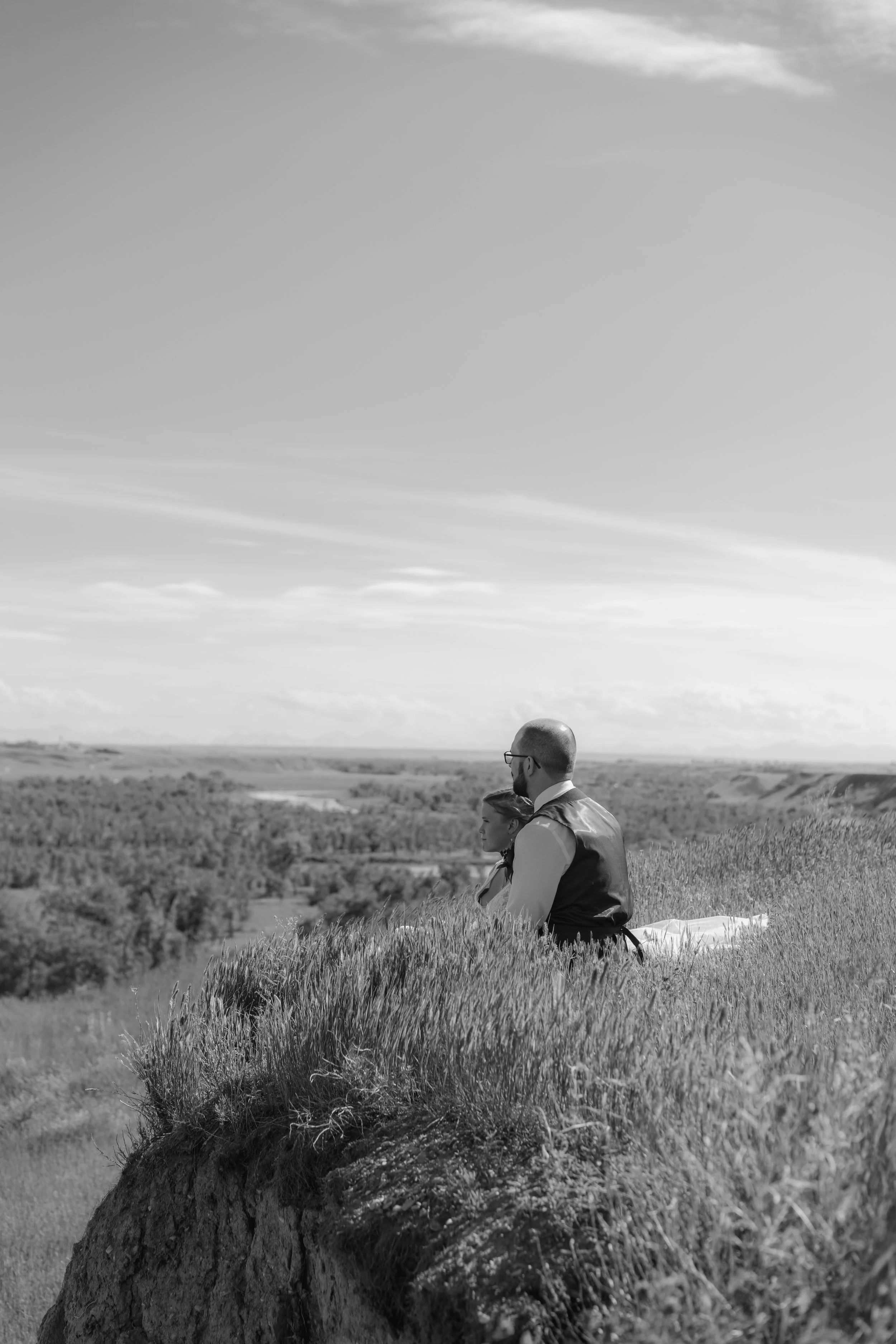 A man and a woman sitting on a grassy hilltop, looking out over a landscape of trees and open land, with a partly cloudy sky above.