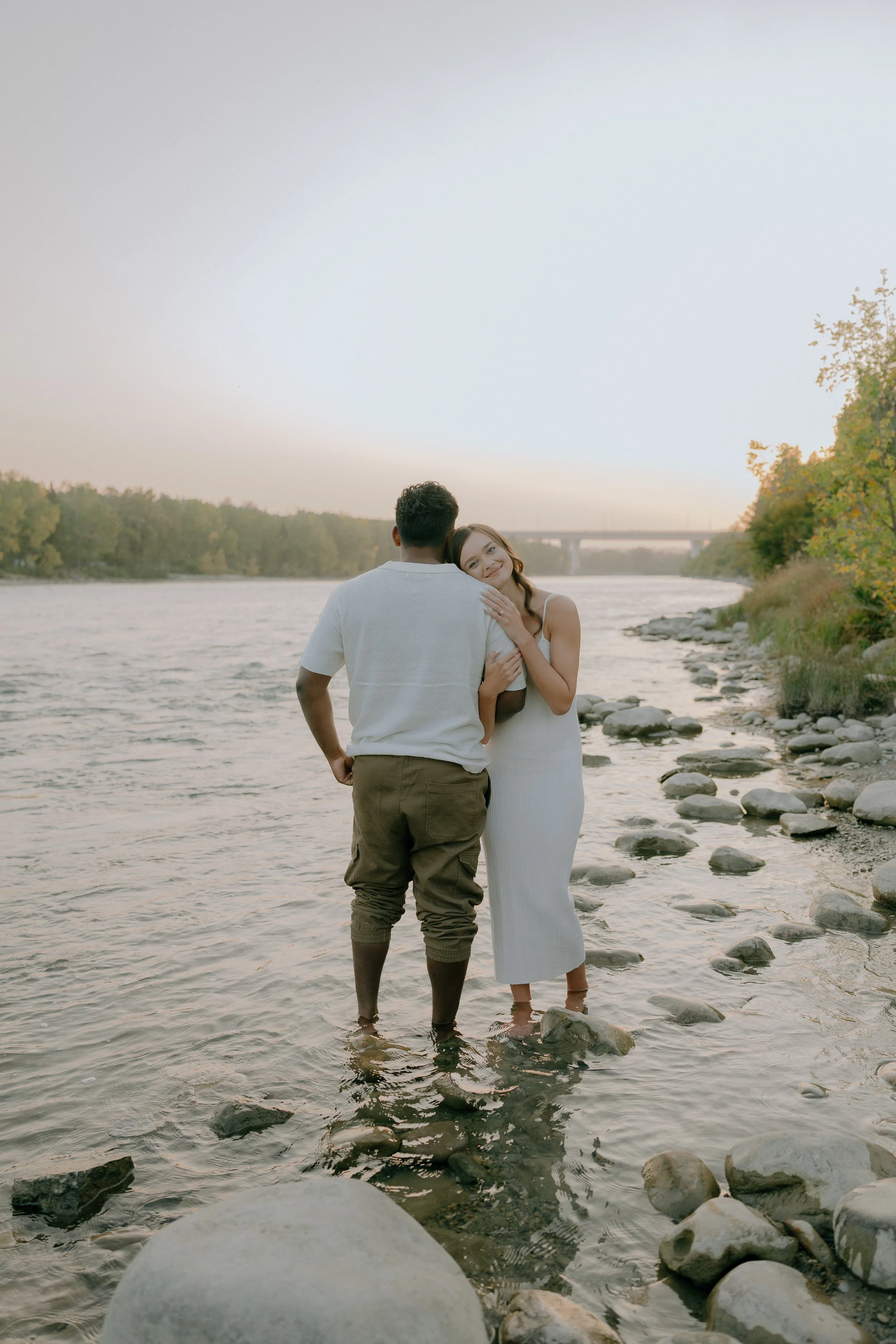 A couple standing in a river at sunset, with the woman leaning her head on the man's shoulder, surrounded by rocks and trees.