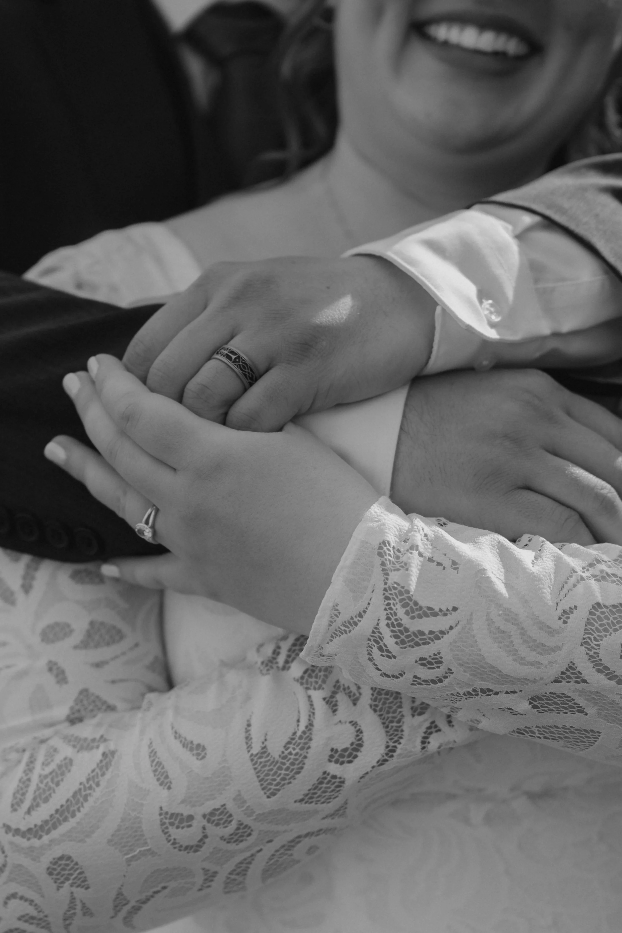 Close-up of a smiling woman wearing a lace wedding dress, holding hands with a person in formal attire, both wearing wedding rings, in a black and white photo.