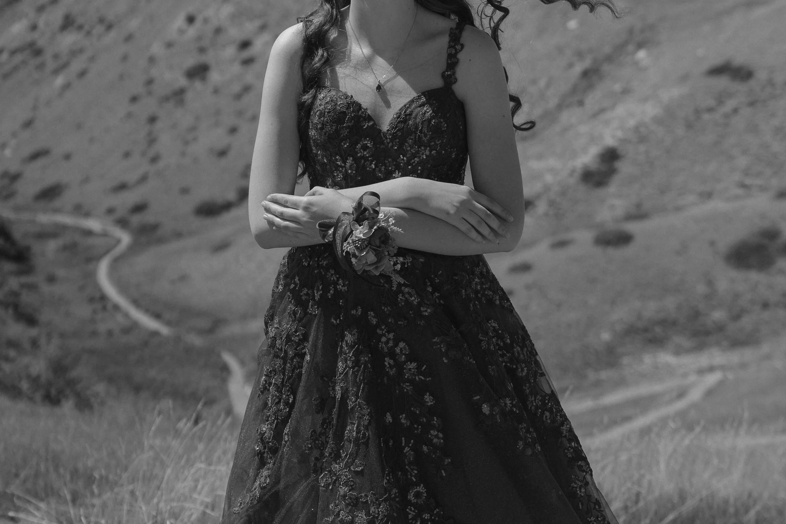 A woman wearing a black lace dress and corsage is standing outdoors on a grassy hill with hills in the background.