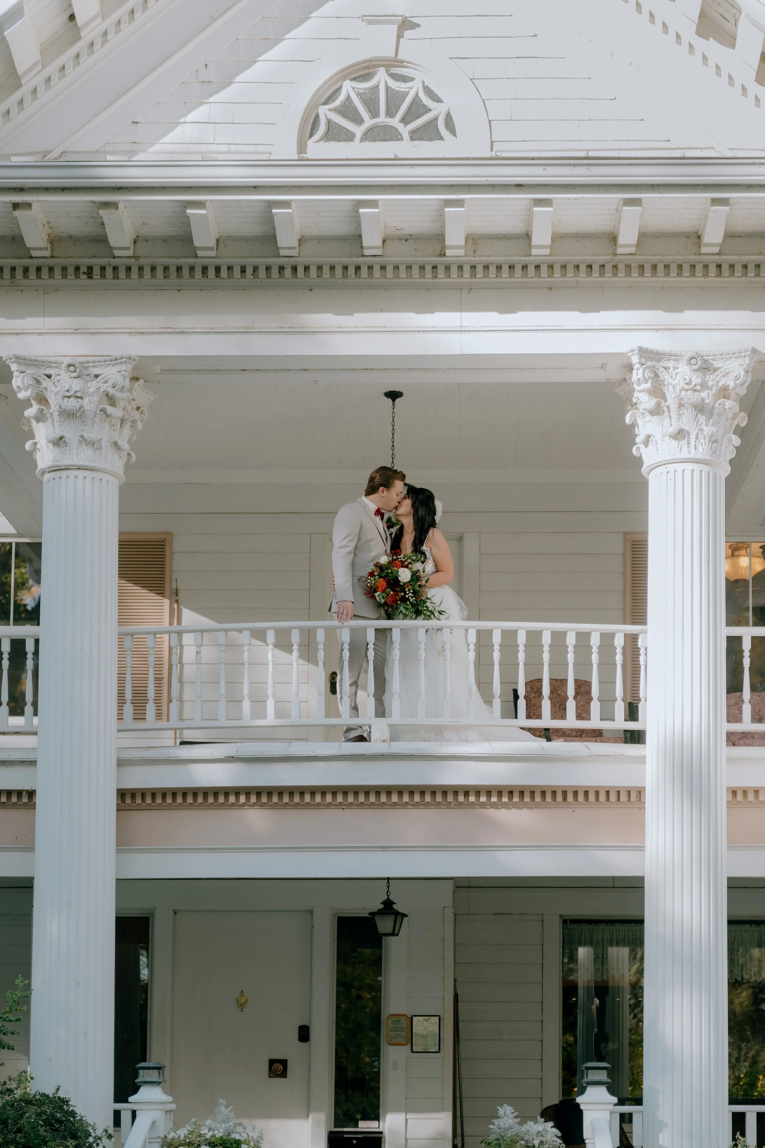 A bride and groom share a kiss on a white porch, with the bride holding a bouquet of flowers, in front of a white house with columns and decorative trim.