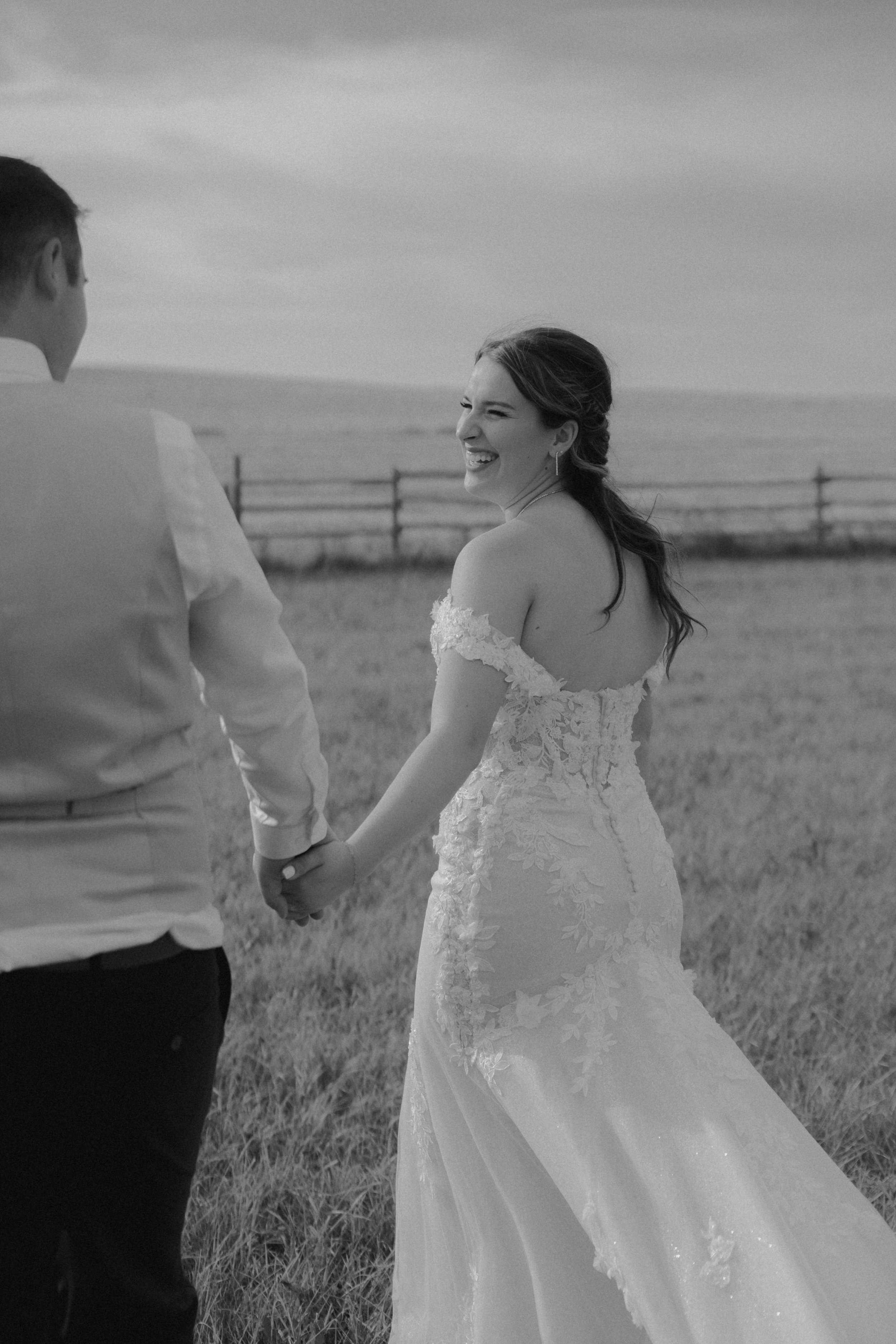 A bride and groom holding hands in a field, smiling at each other, with a wooden fence and open sky in the background.