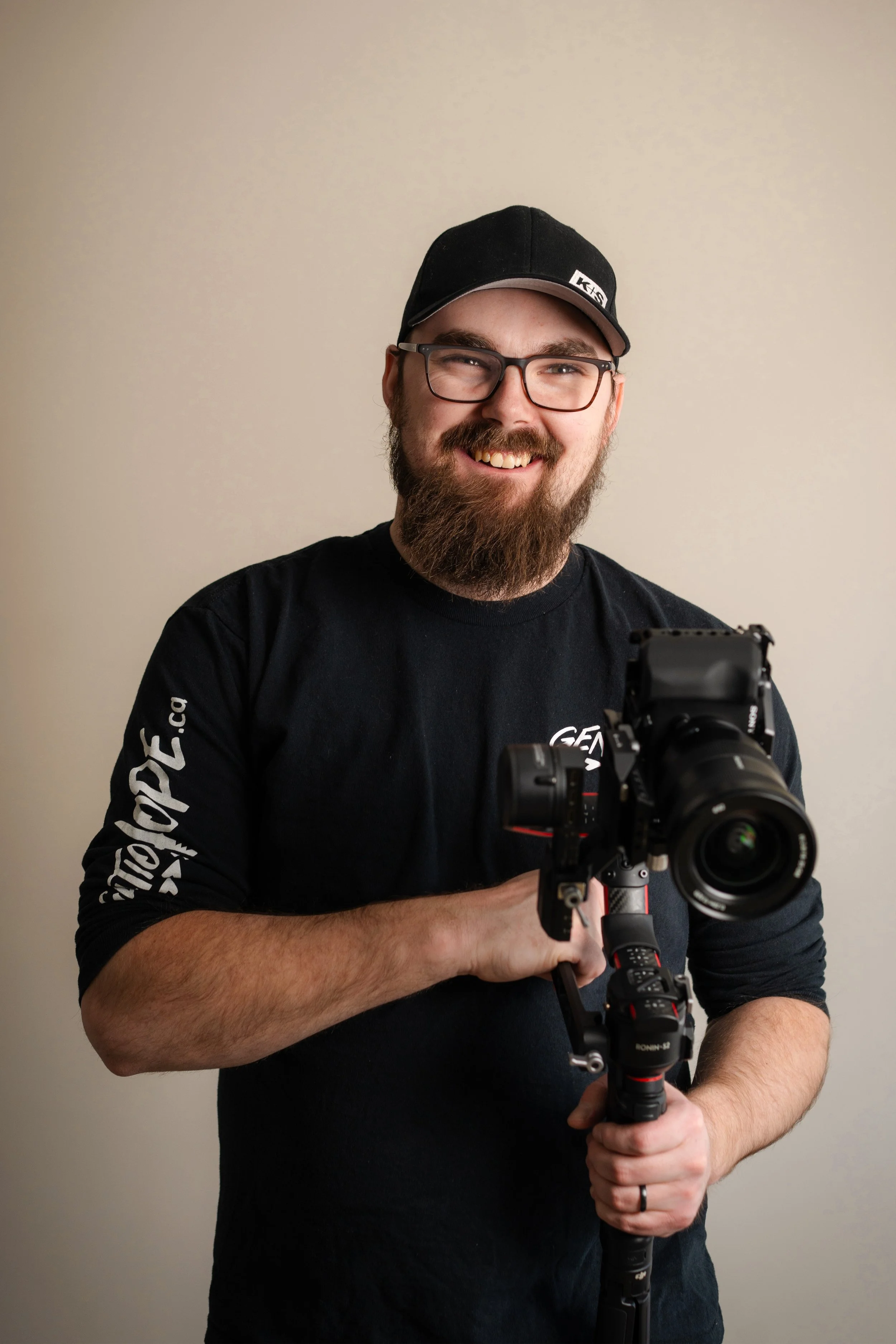 A smiling man with a beard and glasses holding a professional camera on a stabilizer, wearing a black cap and black shirt.