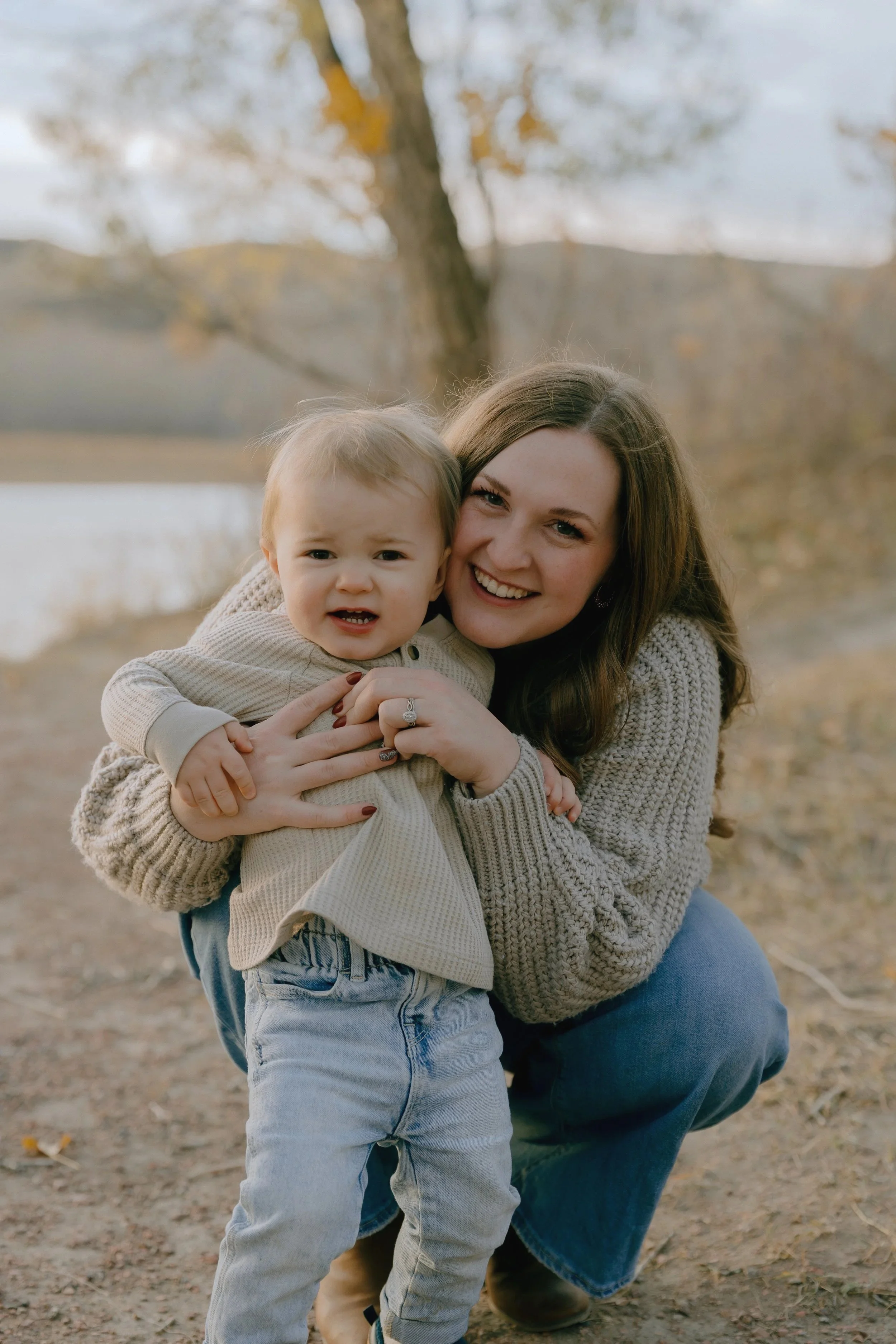 A woman and a young child outdoors near a lake with autumn trees in the background. The woman is smiling and holding the child, who appears confused or concerned.