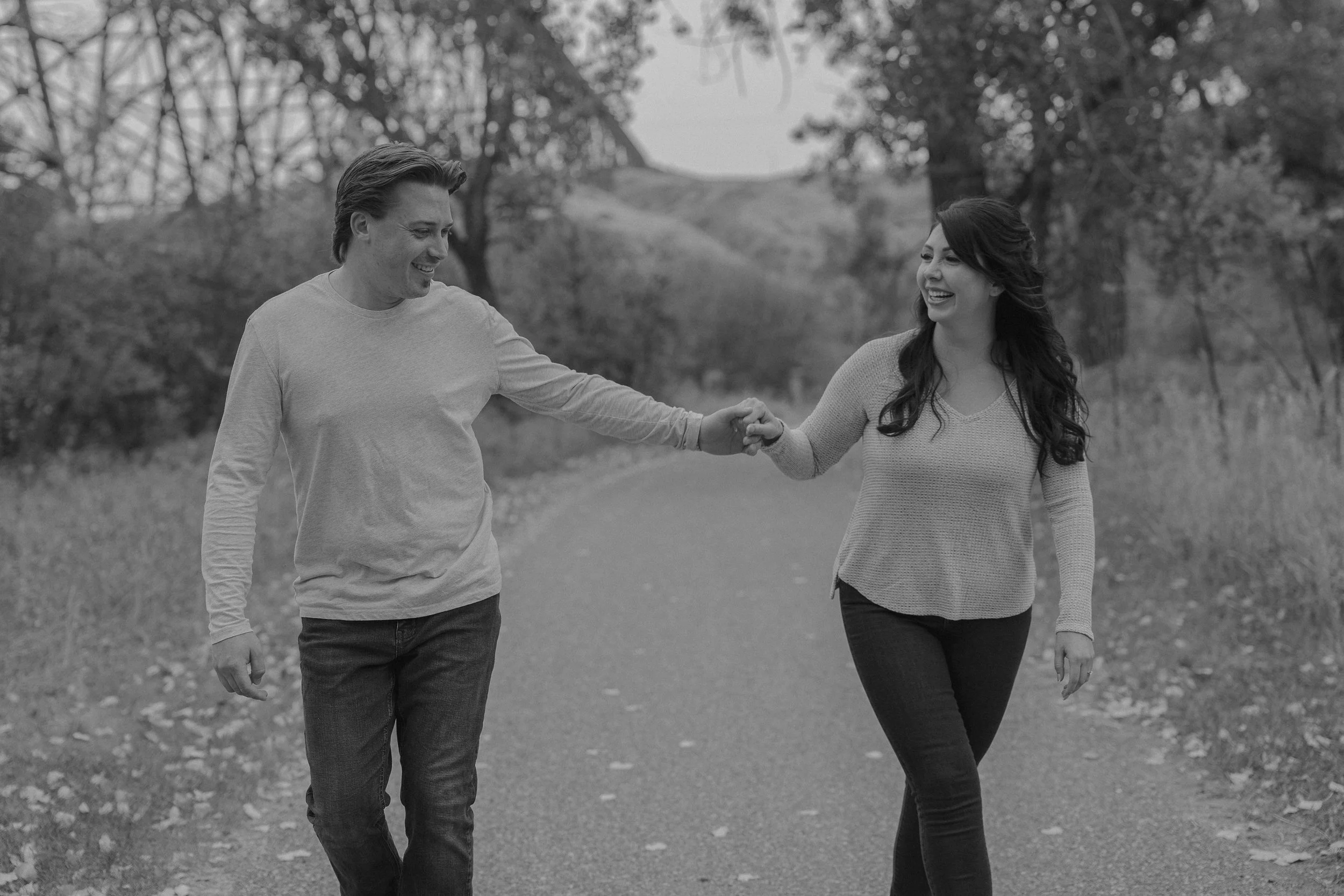 A man and woman walking outdoors on a path, holding hands and smiling at each other, surrounded by trees and natural scenery.
