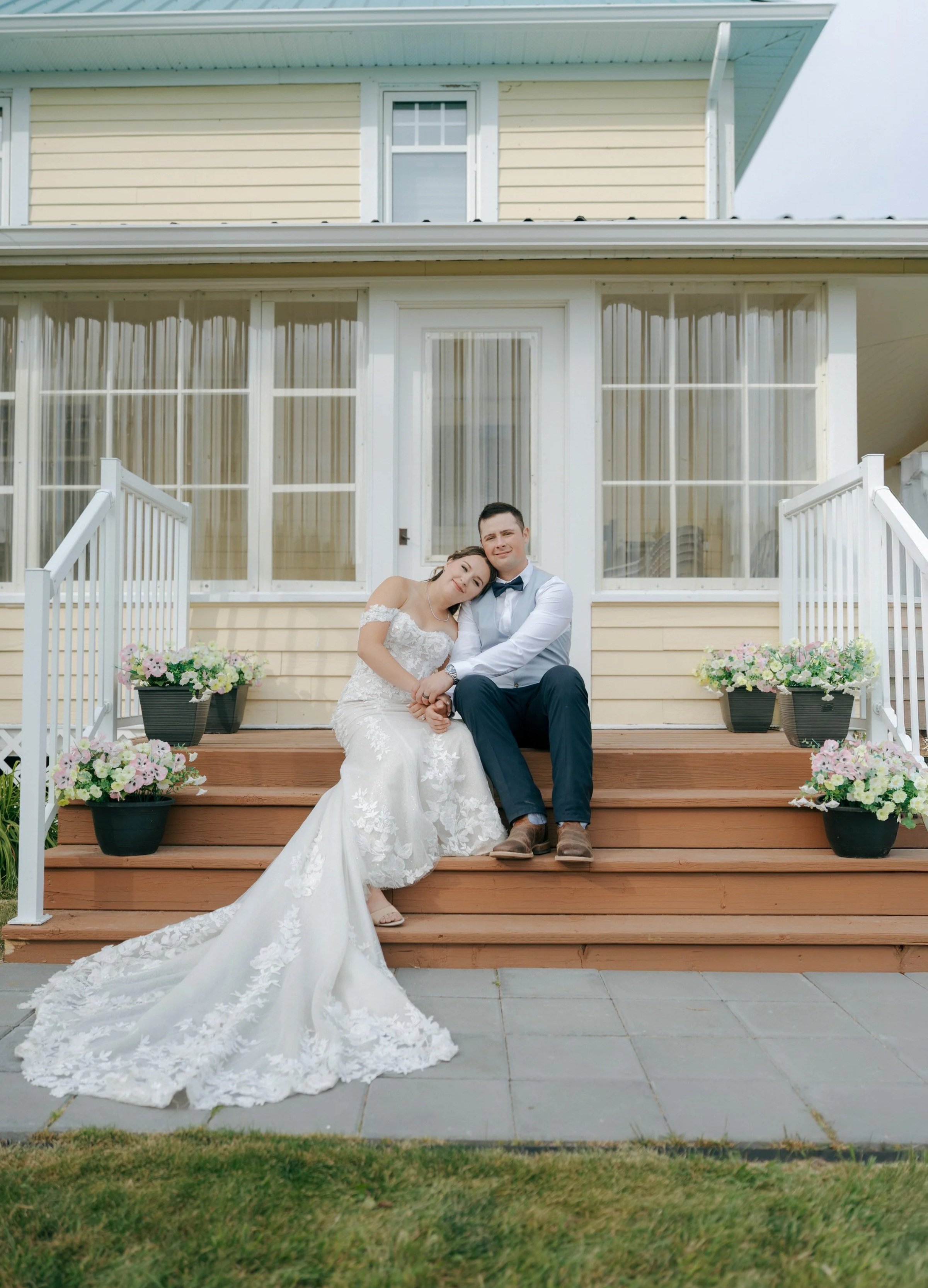 A newlywed couple sitting on the steps of a yellow house, with the bride resting her head on the groom's shoulder. The bride is wearing a white lace wedding gown, and the groom is in a white shirt with a bow tie and dark pants. Four flower pots with pink and white flowers decorate the steps.