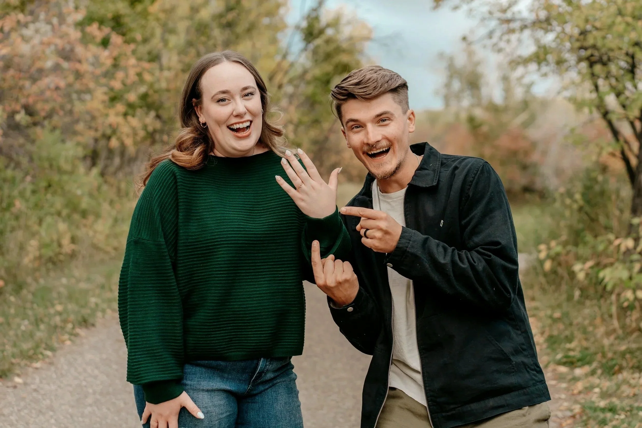 A happy couple standing outdoors on a pathway surrounded by trees with fall foliage, showing off an engagement ring.