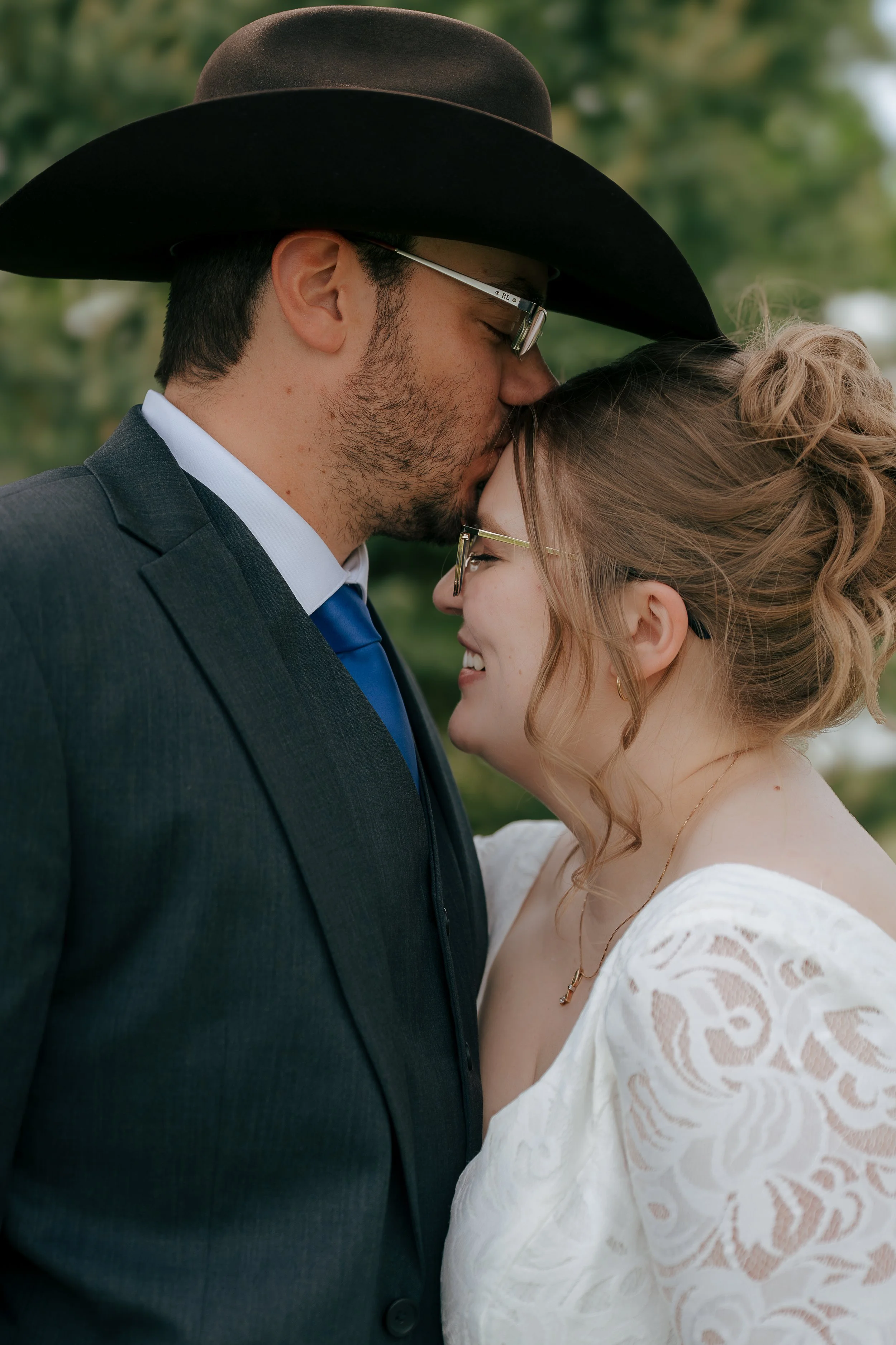 A couple dressed in formal attire, with the man wearing a suit and a cowboy hat, and the woman wearing a white dress, sharing an intimate moment outdoors with a blurred green background.