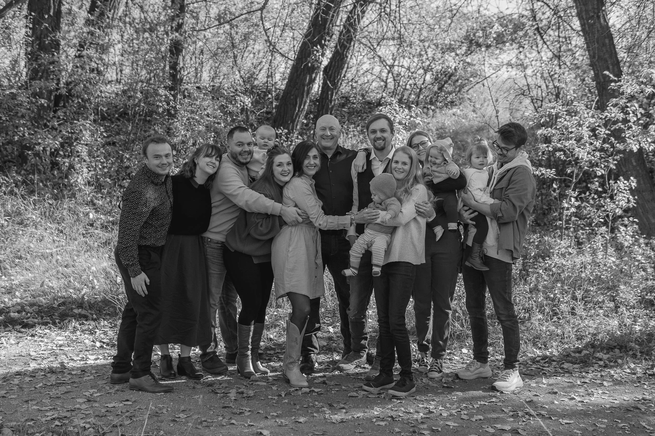 A large group of people, including adults and children, standing outdoors in a wooded area, smiling and hugging each other.