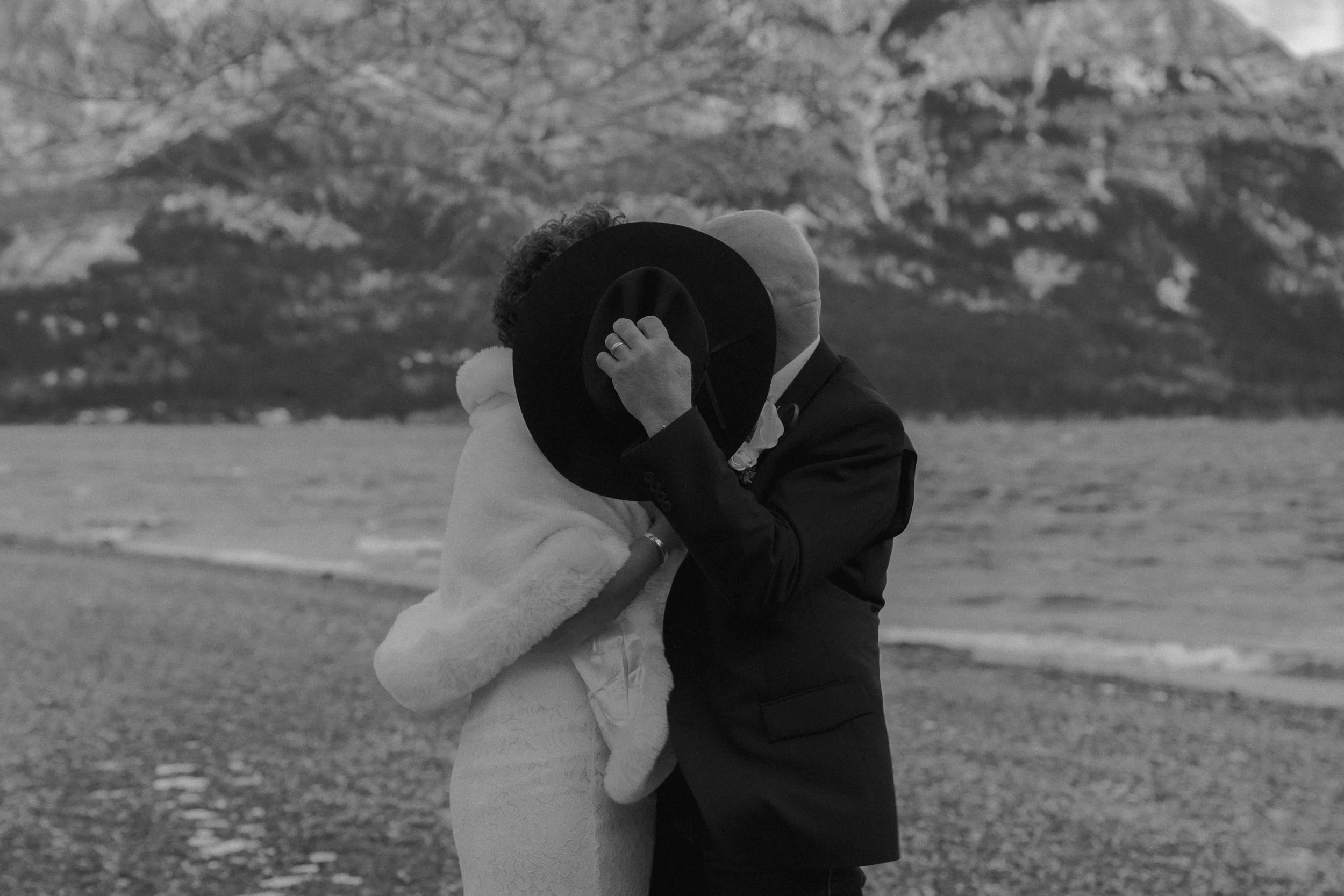 A couple sharing an intimate moment on a beach, with a mountain in the background, both holding a large hat to their faces.