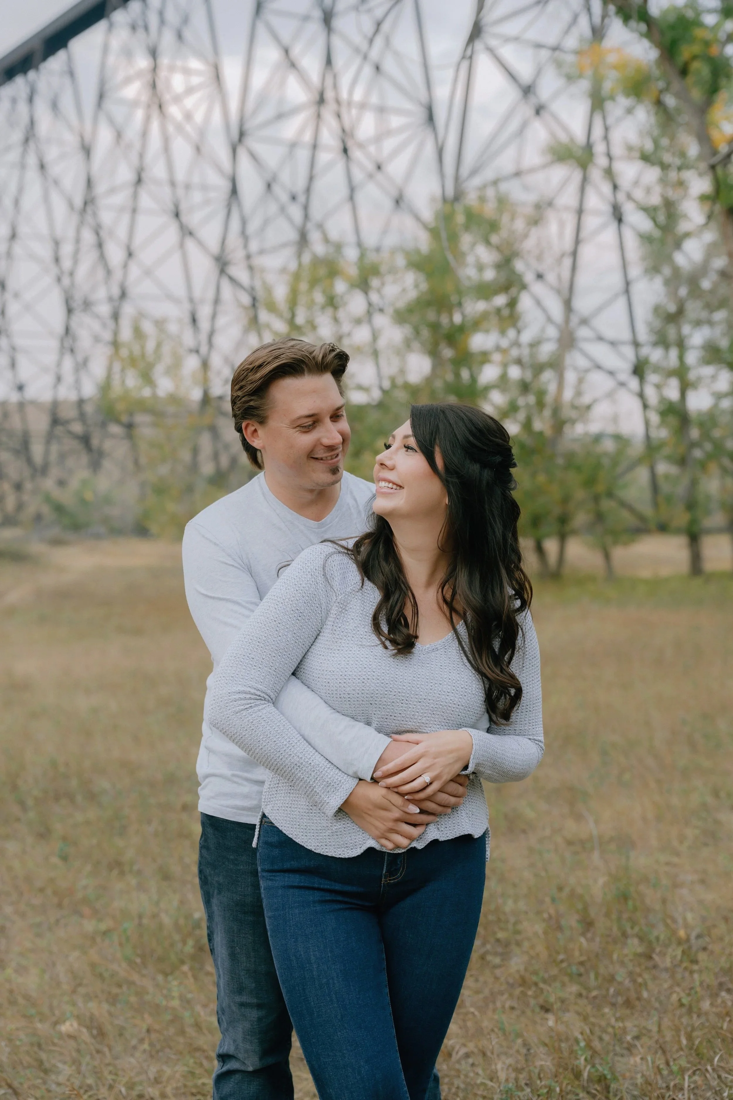 A couple standing outdoors in a grassy field, smiling and embracing each other with a metal power line tower in the background.