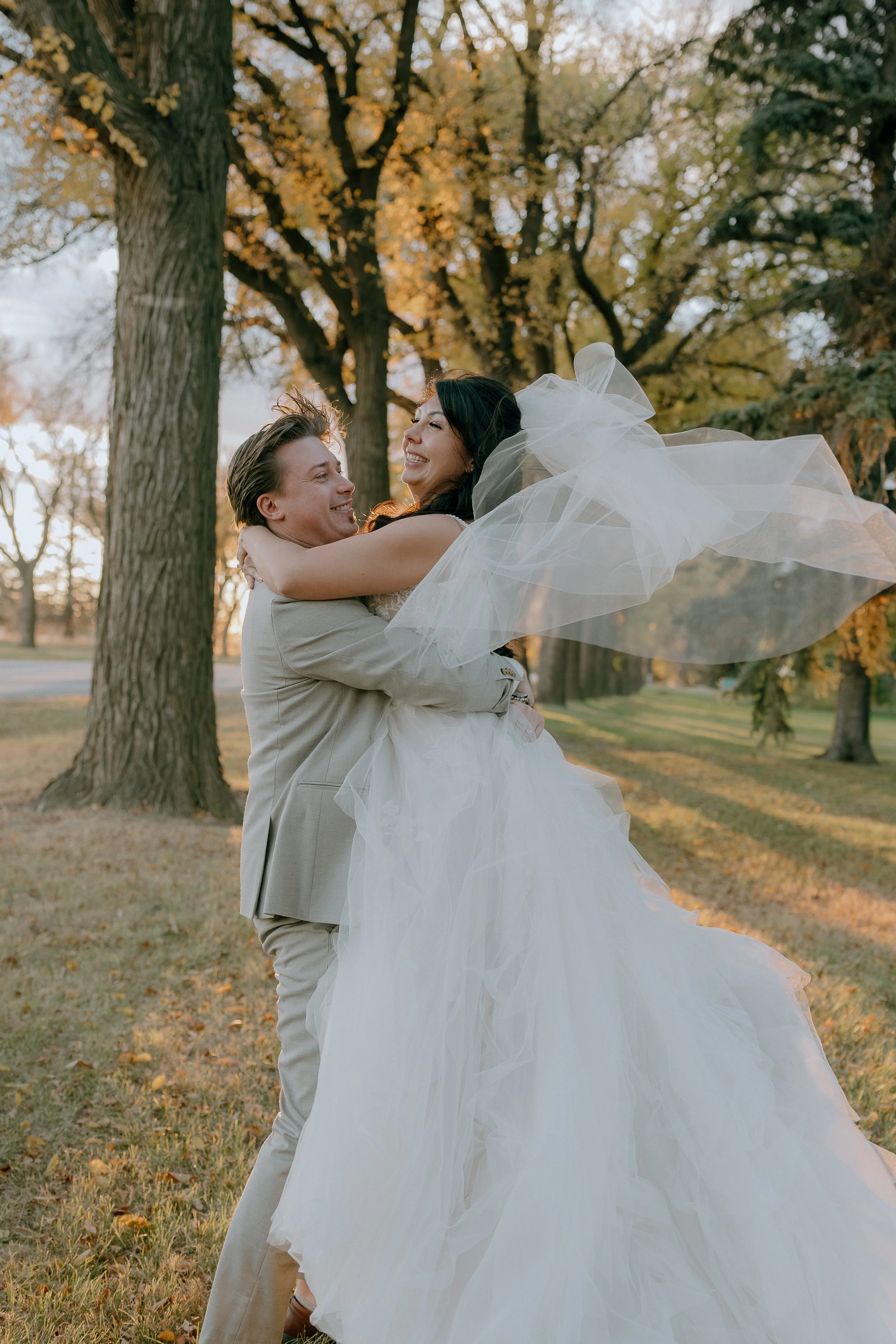 A bride with a long white wedding gown and veil being lifted in the air by a groom in a light-colored suit, smiling and embracing each other in a park with trees and autumn leaves.