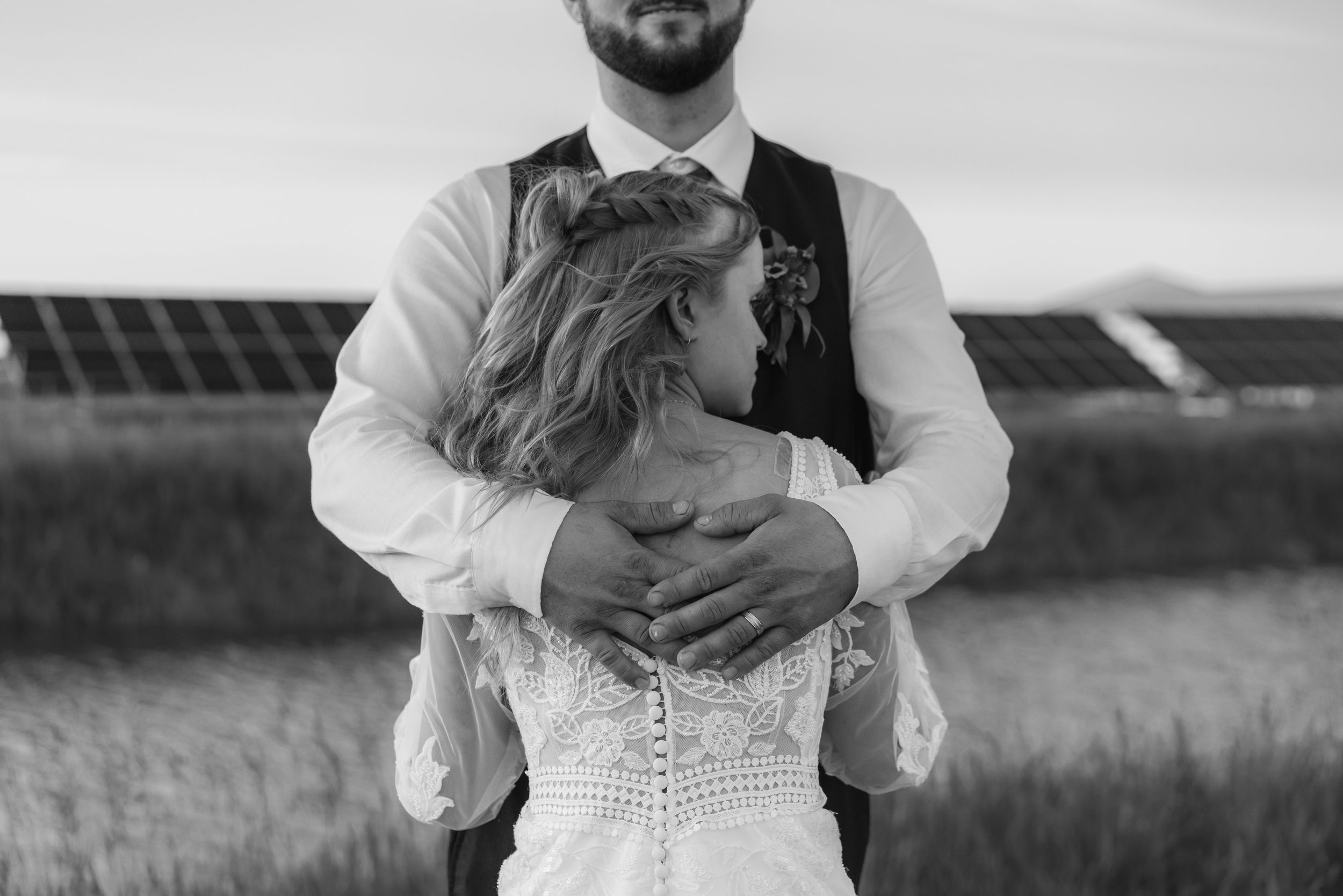 A couple embracing outdoors, with the woman wearing a lace wedding dress and the man in a dress shirt and vest, in a field with solar panels in the background, black and white photograph.