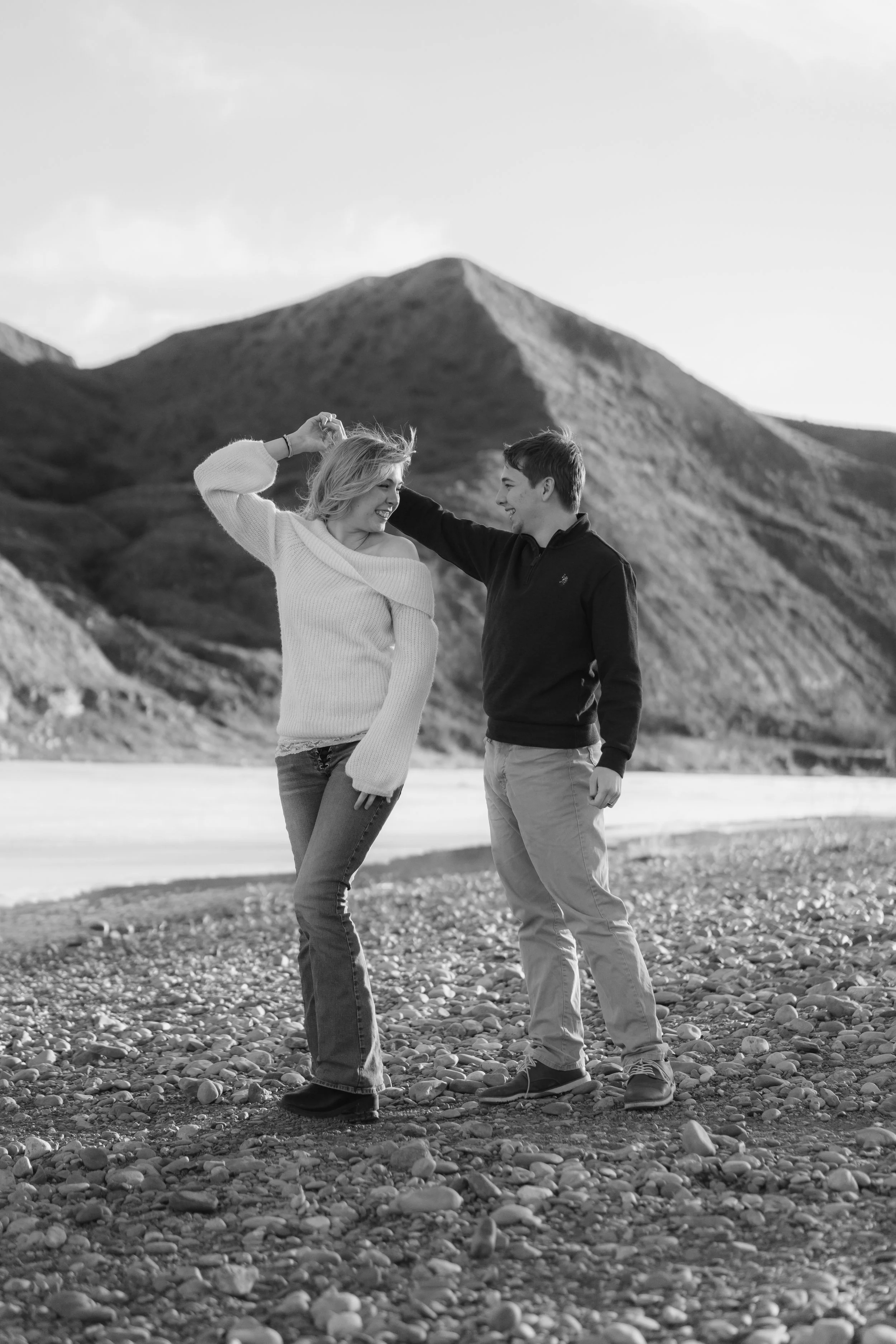 A black-and-white photo of a smiling young couple dancing on a rocky beach with mountains in the background.