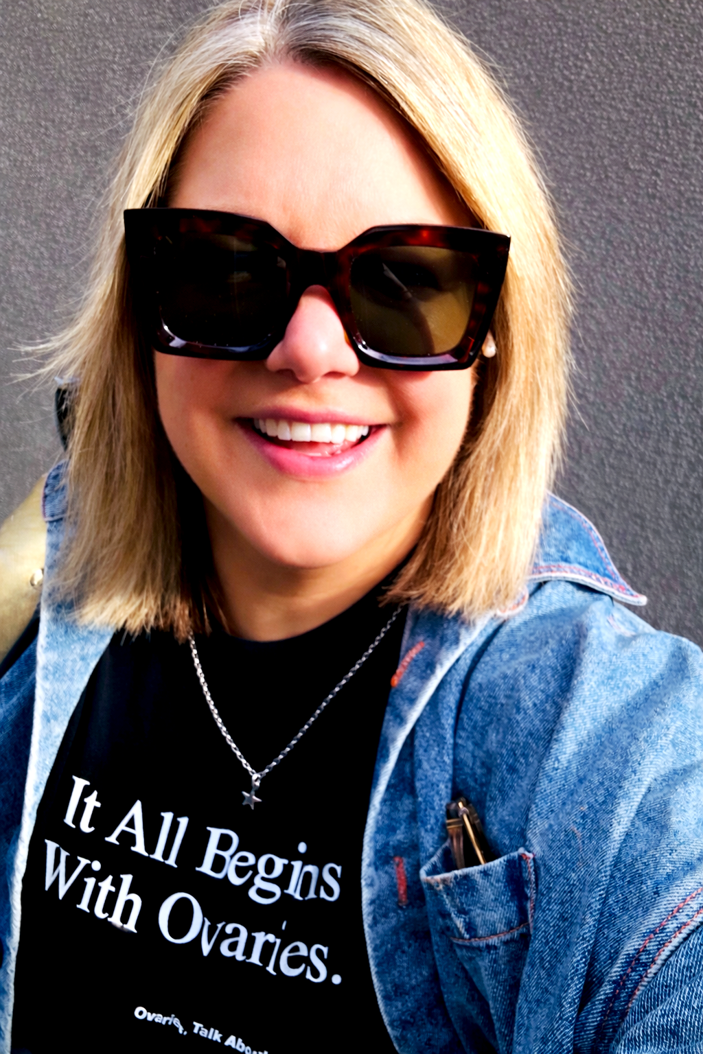 A woman smiling wearing large dark sunglasses and a denim jacket with a black shirt that reads, 'It All Begins With Ovaries.' She has a necklace with a star pendant and is standing against a gray background.