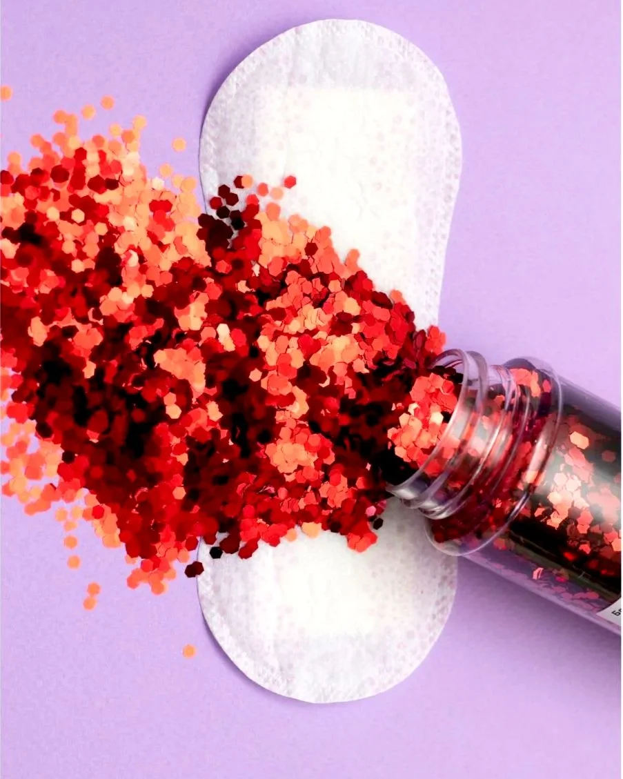Red, pink, black, and orange hexagonal glitter spilling from a clear plastic jar onto a purple surface with a white textured oval paper underneath.