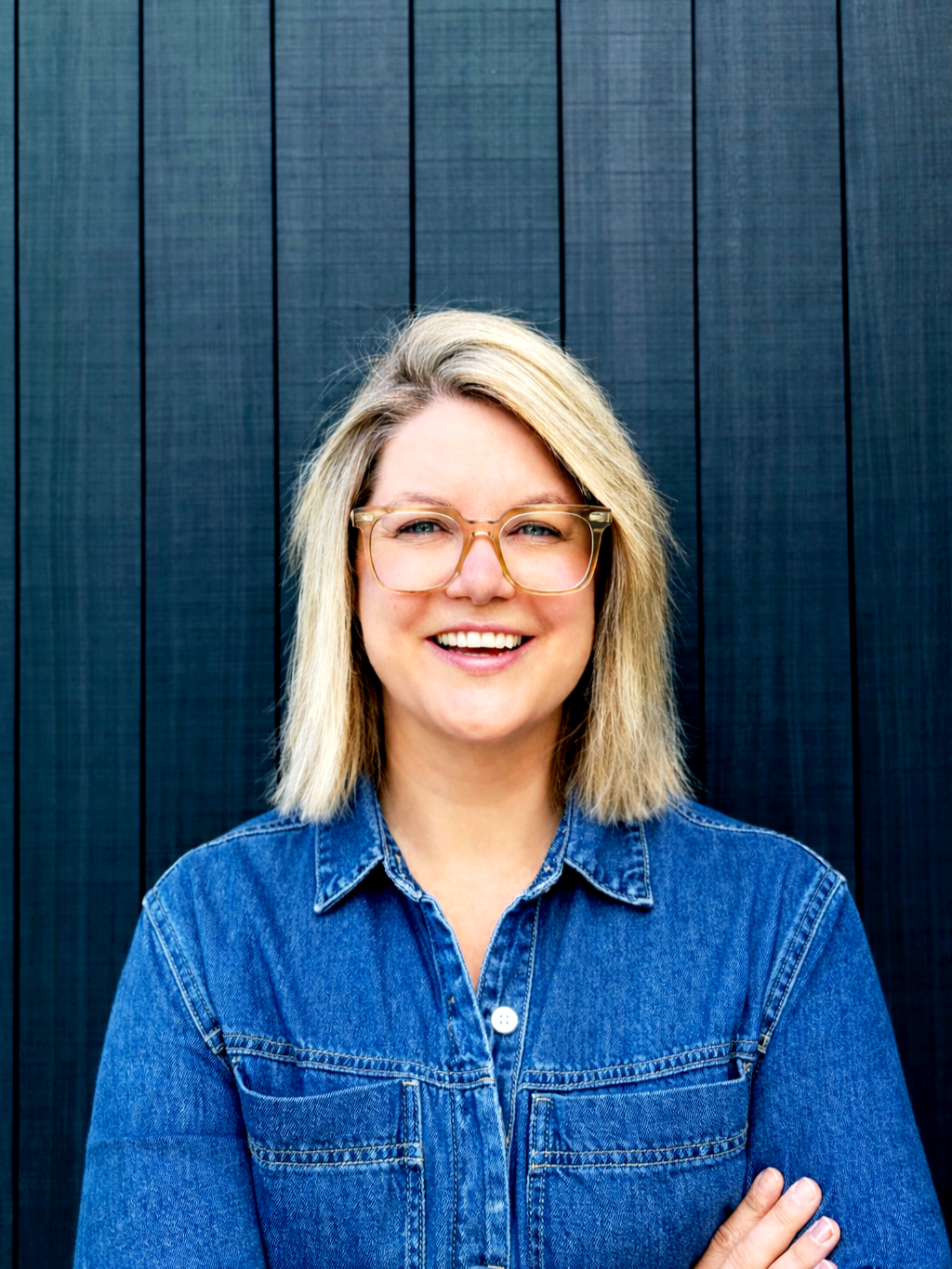 Smiling woman with blonde hair, wearing glasses and a denim jacket, standing in front of a dark wooden background.