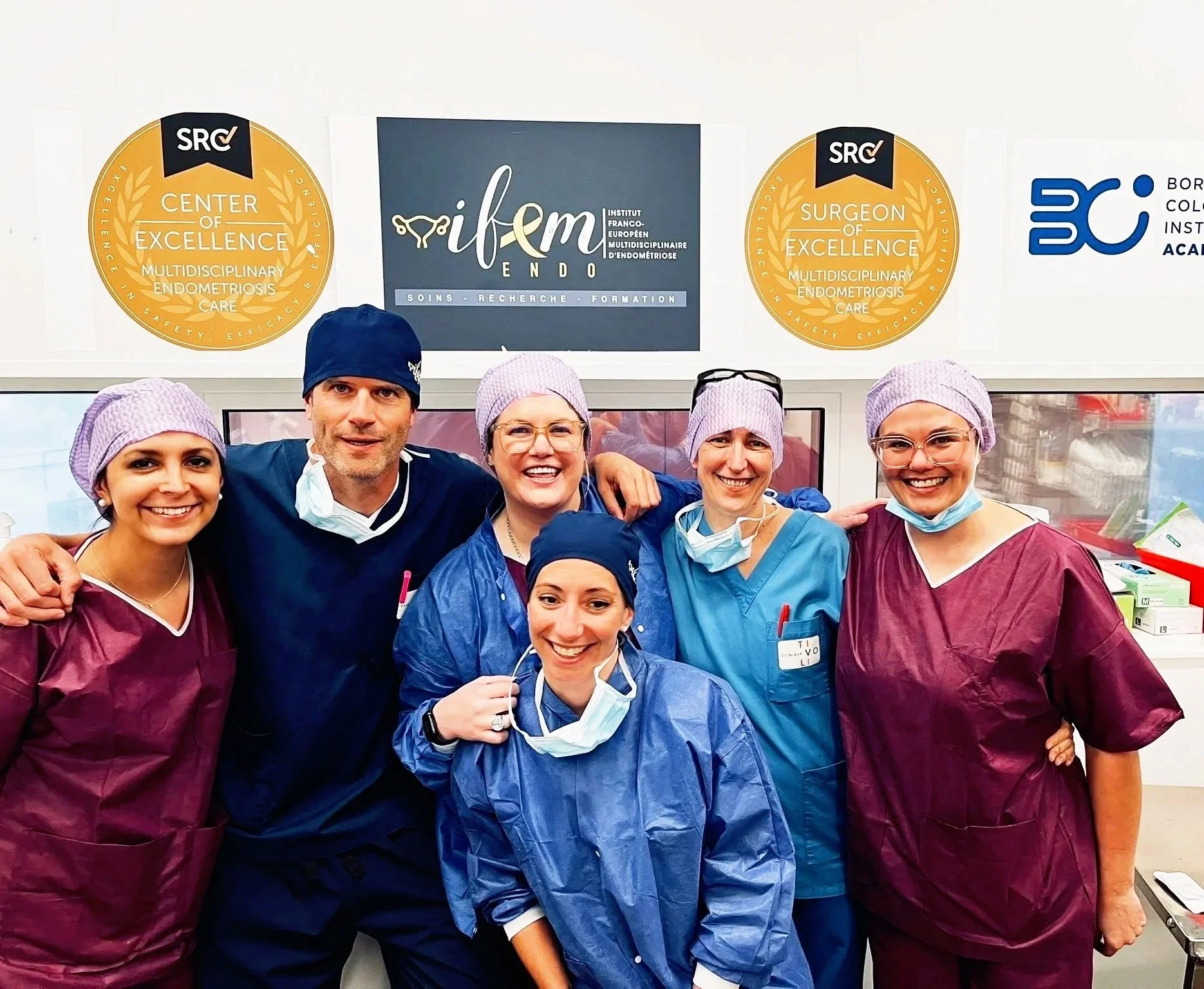 Group of medical professionals wearing scrubs and surgical caps, smiling and posing together in a hospital or clinic setting.