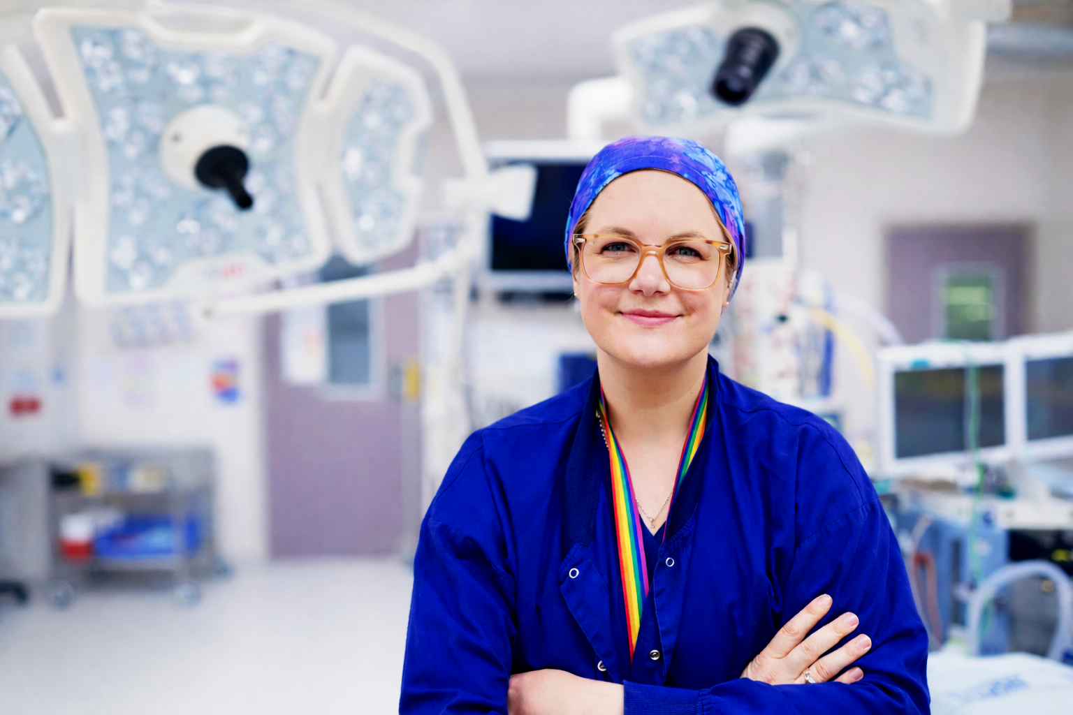 A smiling healthcare professional in scrubs and glasses in a medical setting with surgical lights and equipment in the background.