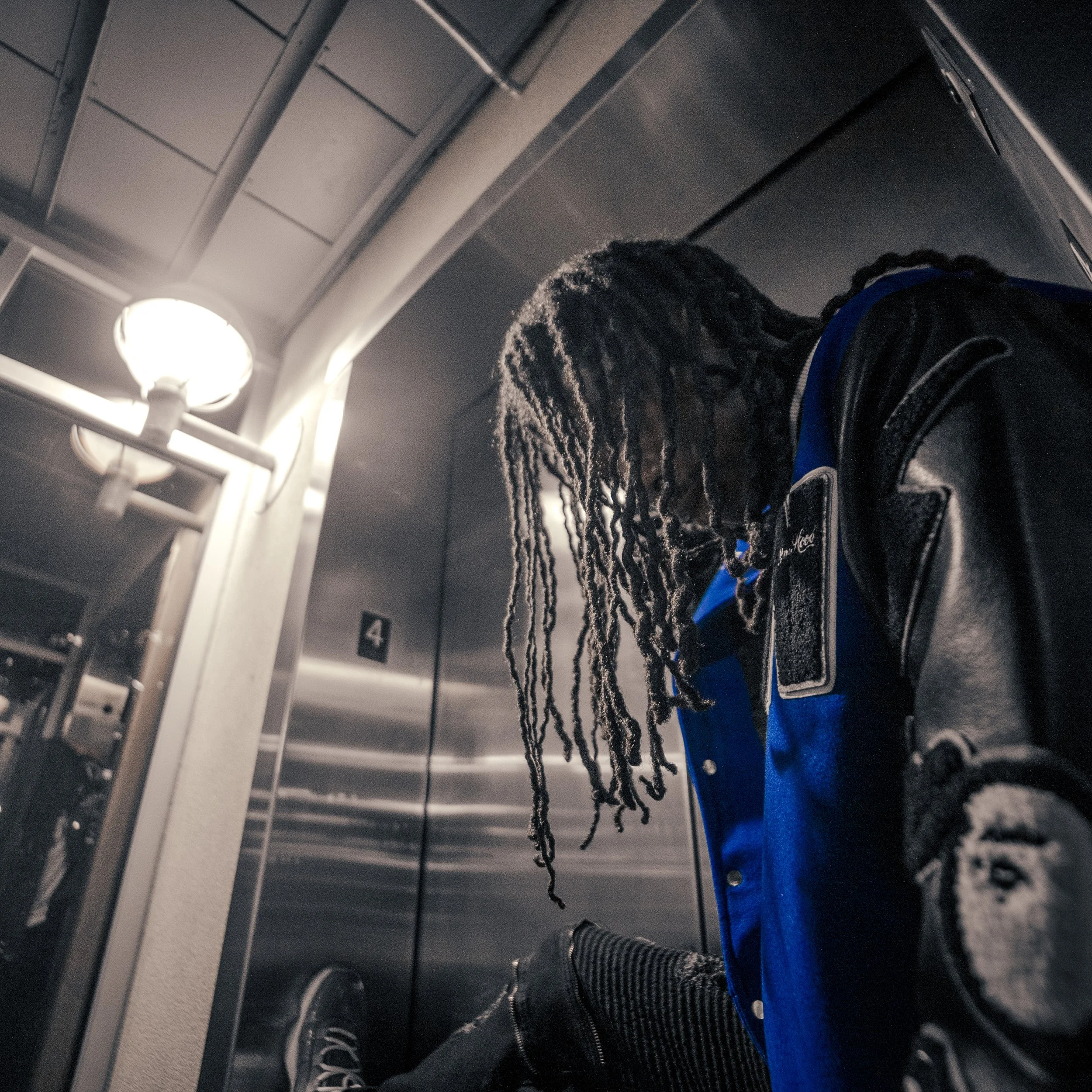 A person with dreadlocks wearing a black and blue varsity jacket, sitting in an elevator with metallic walls, looking down.