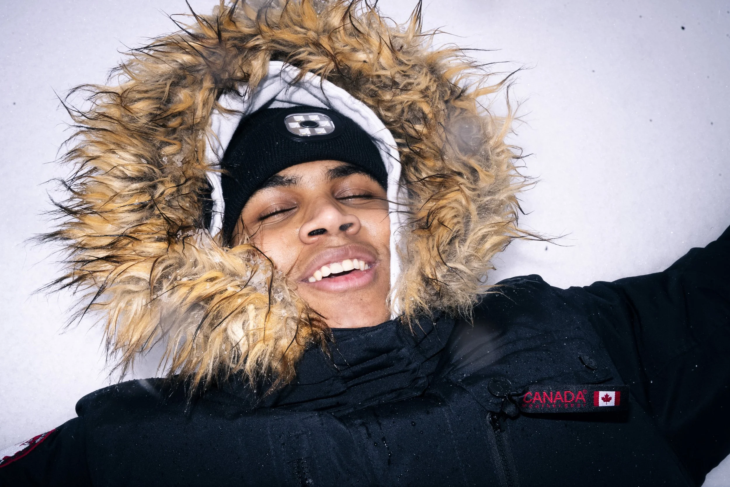 Young man lying on snow with a fur-lined hood, wearing a black Canada-themed jacket and beanie, smiling with eyes closed.