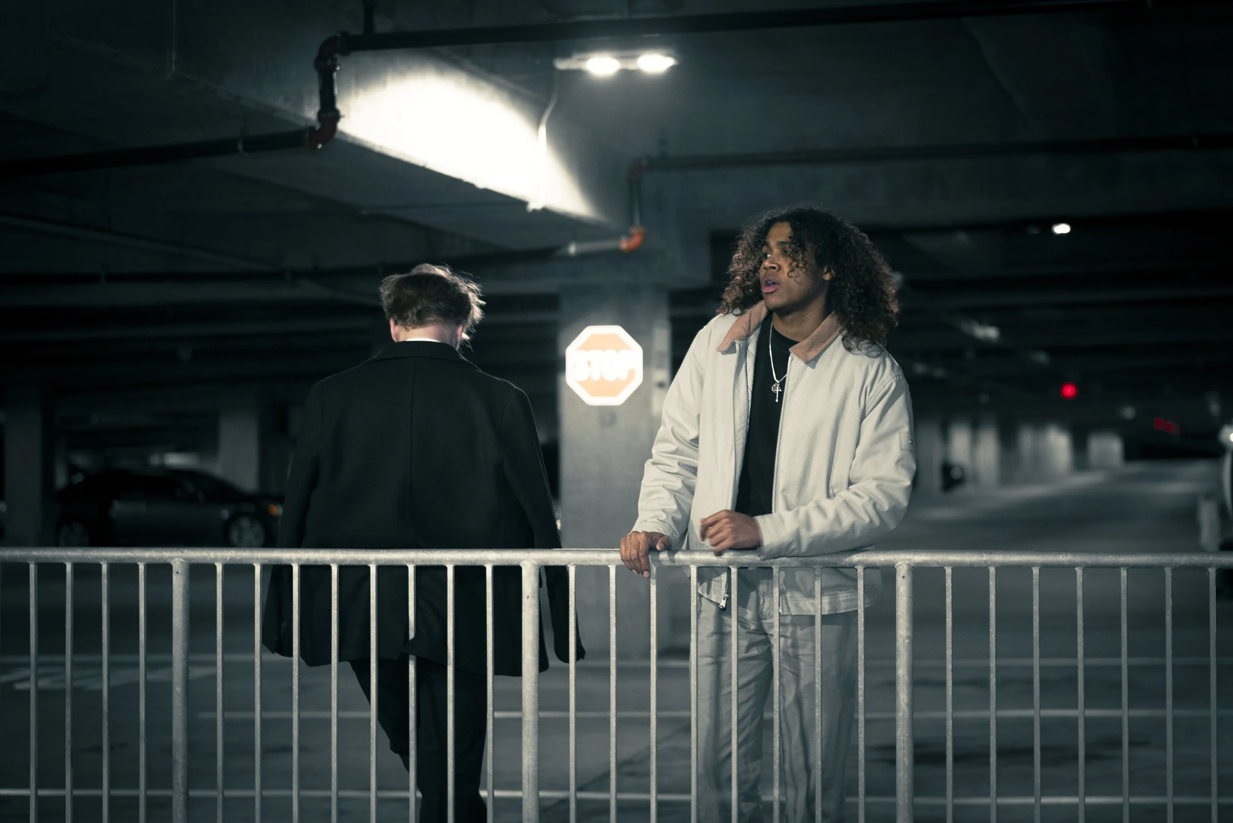 A man with curly hair in a beige jacket leaning on a railing in a parking garage, with a woman in black facing away, walking away in the background.
