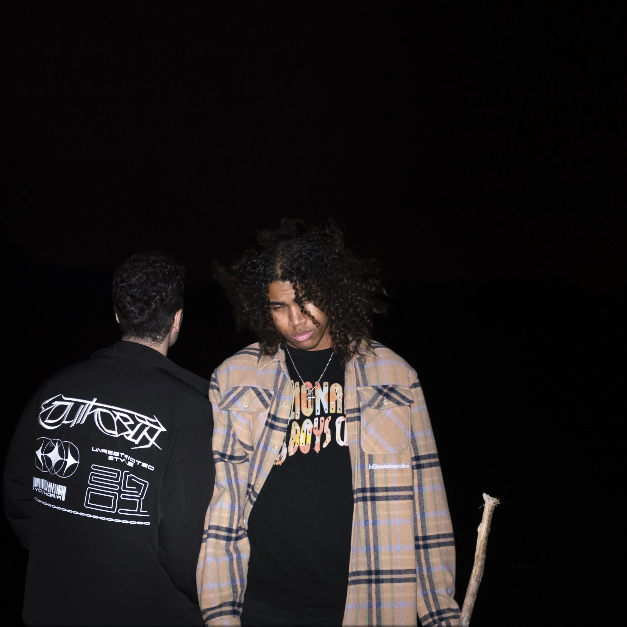 A woman with big, curly hair wearing a plaid jacket and a black graphic t-shirt, standing with her eyes closed, and a man with short dark hair in a black jacket with white text, facing away, against a dark background.