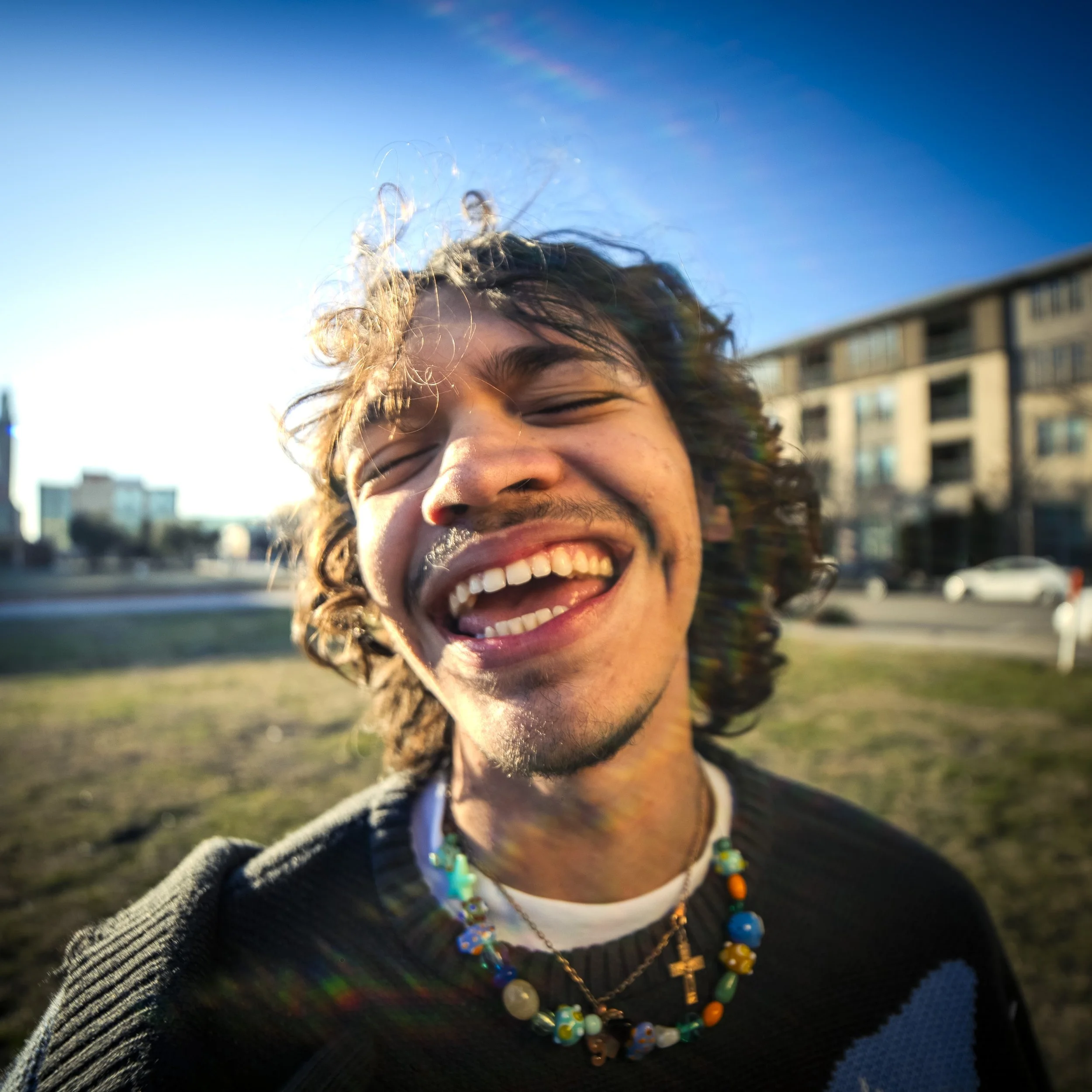 A young man with curly hair smiling broadly outdoors during the daytime, wearing multiple necklaces including one with a cross, against a background of buildings and cars.