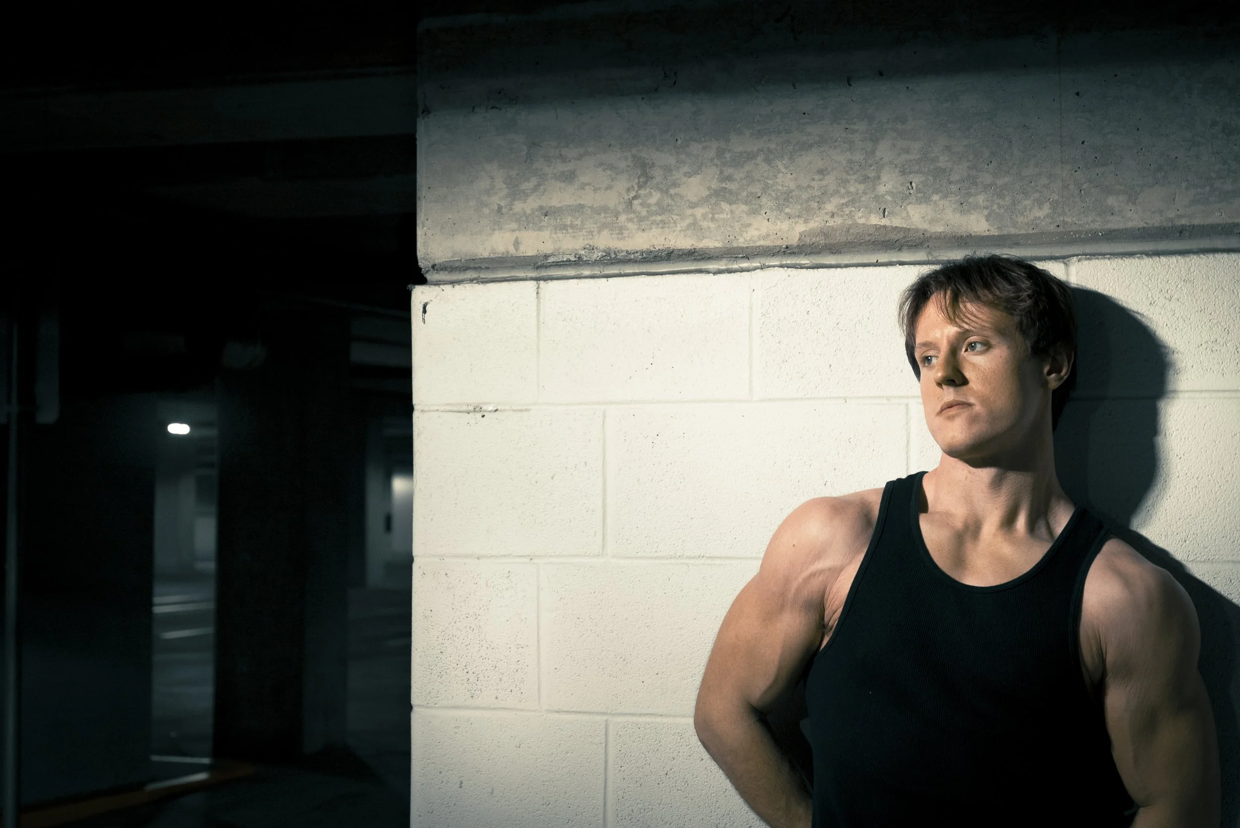 A man with short brown hair, wearing a black sleeveless shirt, leans against a white cinderblock wall in a dimly lit parking garage.