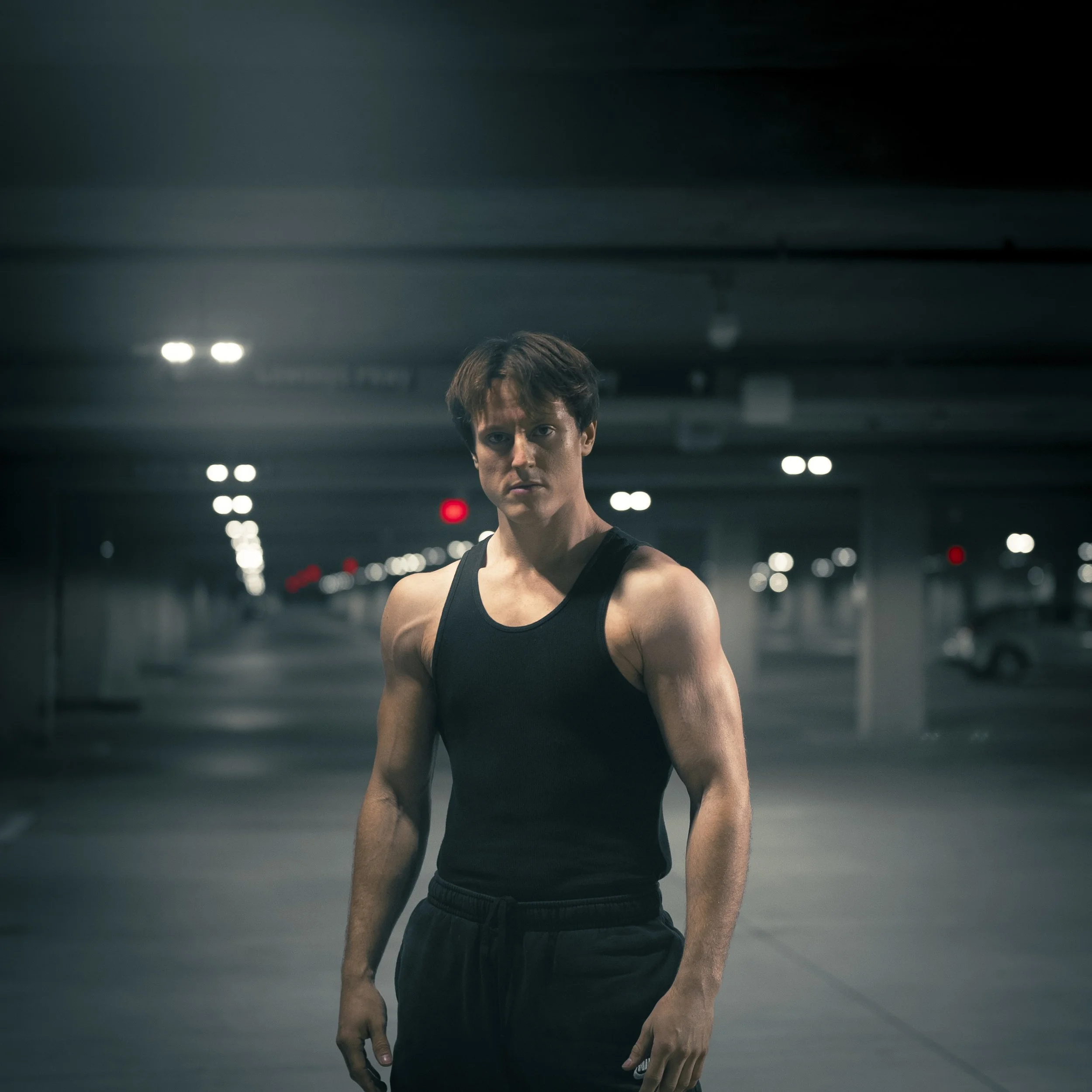 A young man with short brown hair wearing a black sleeveless athletic shirt and black pants, standing in an underground parking lot with dim lighting and cars in the background.
