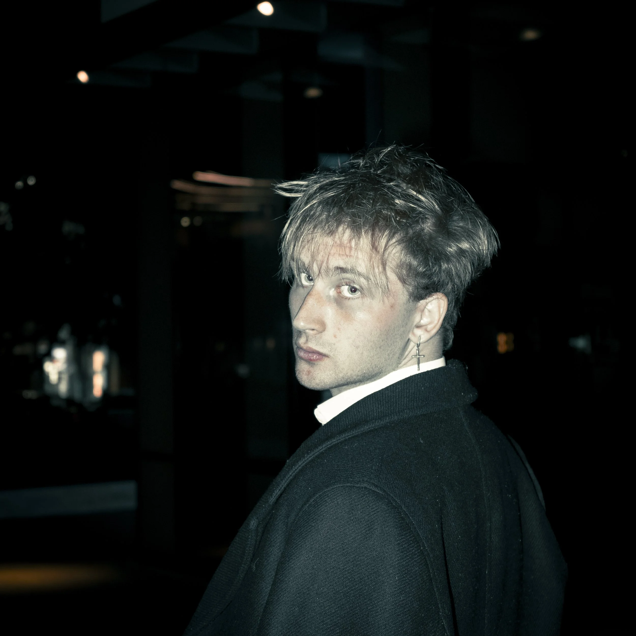 A young man with tousled hair and light skin, wearing a dark coat and a white shirt, looking over his shoulder in a dimly lit environment, with a cross earring visible in his left ear.