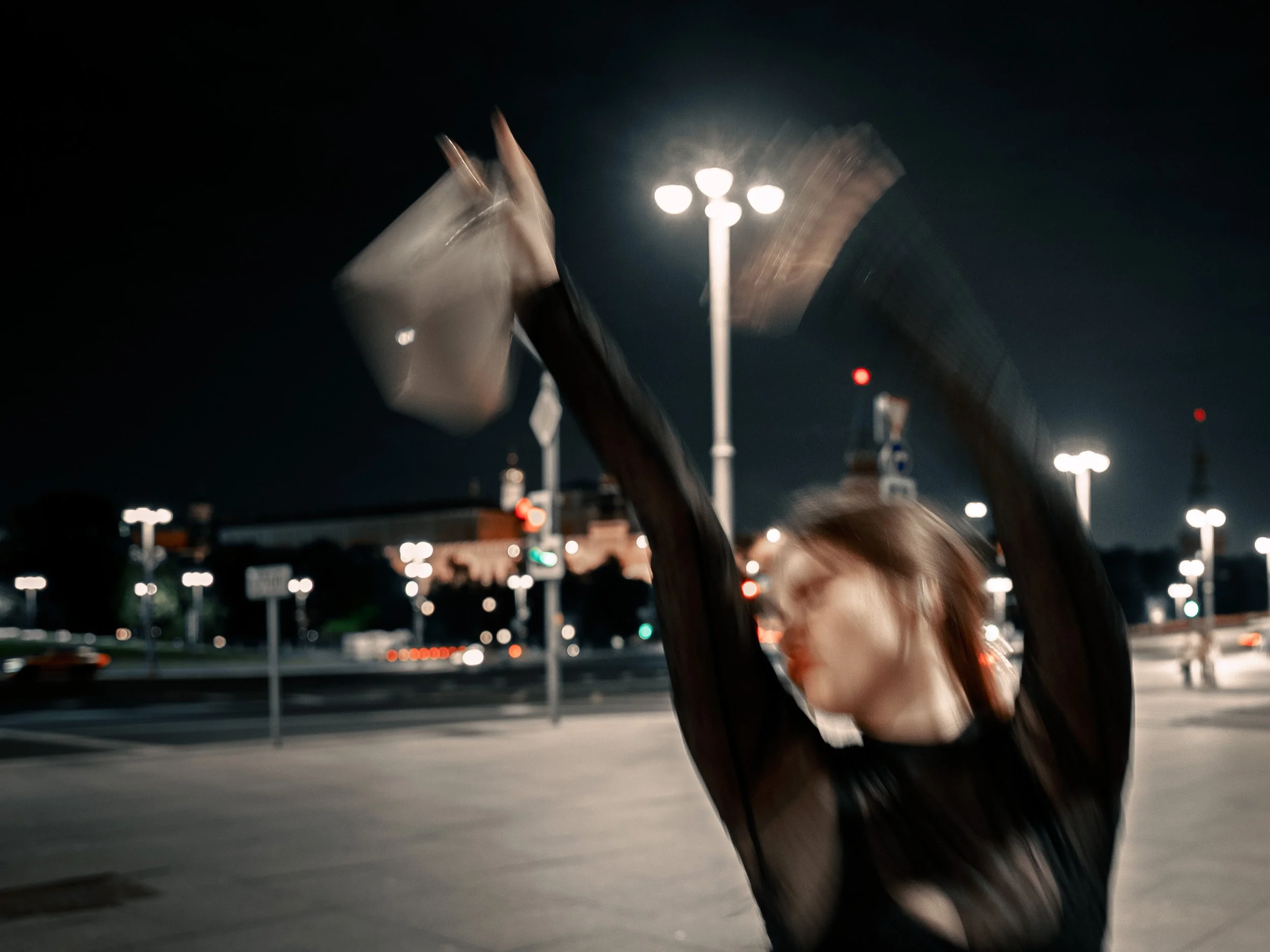 Blurry photo of a woman dancing outdoors at night, with her arms raised, under streetlights, background includes cars and traffic lights.
