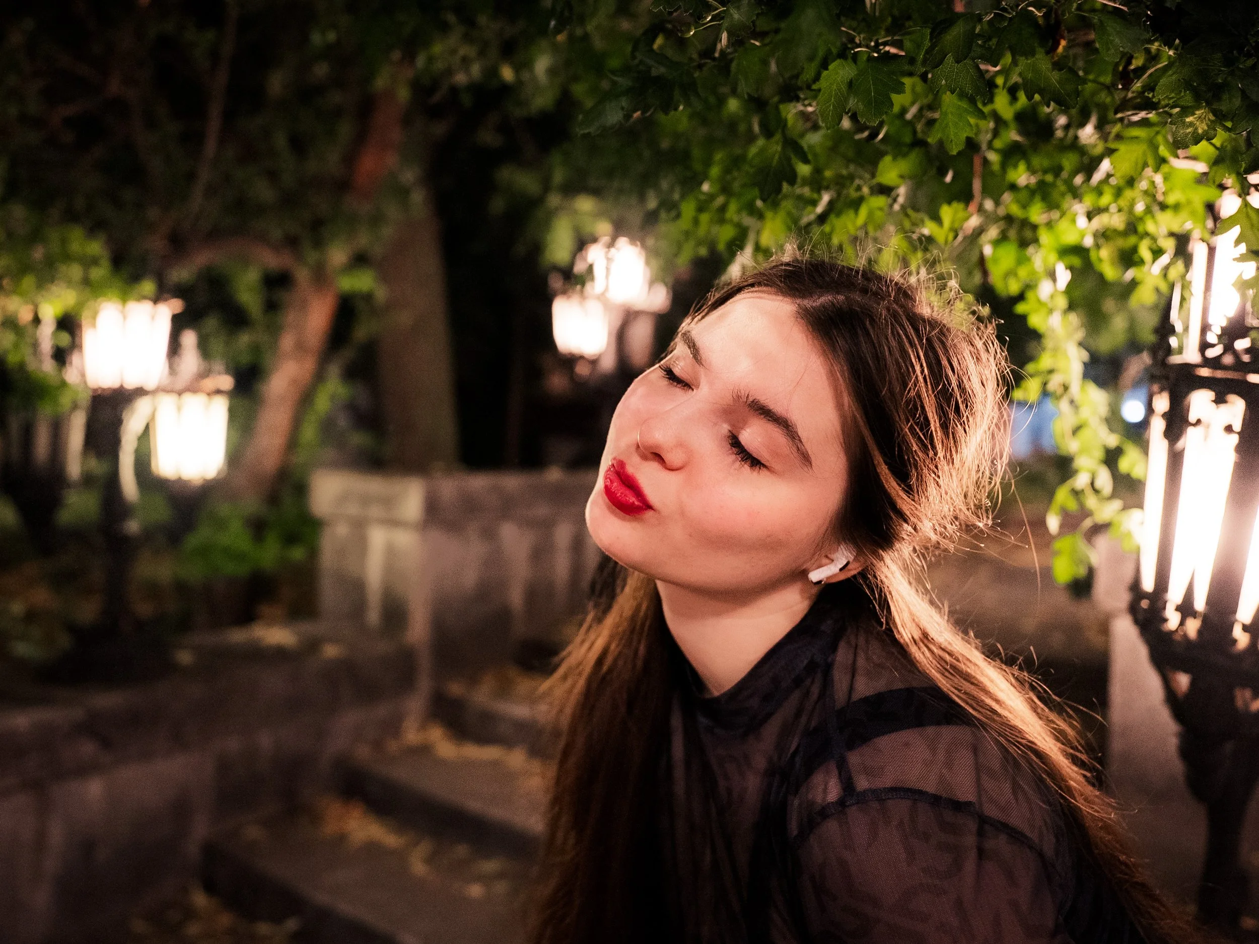 A young woman with long hair and red lipstick is outdoors at night, with her eyes closed and a relaxed expression. She is wearing black clothing and a white earbud, and there are trees and lit lanterns in the background.