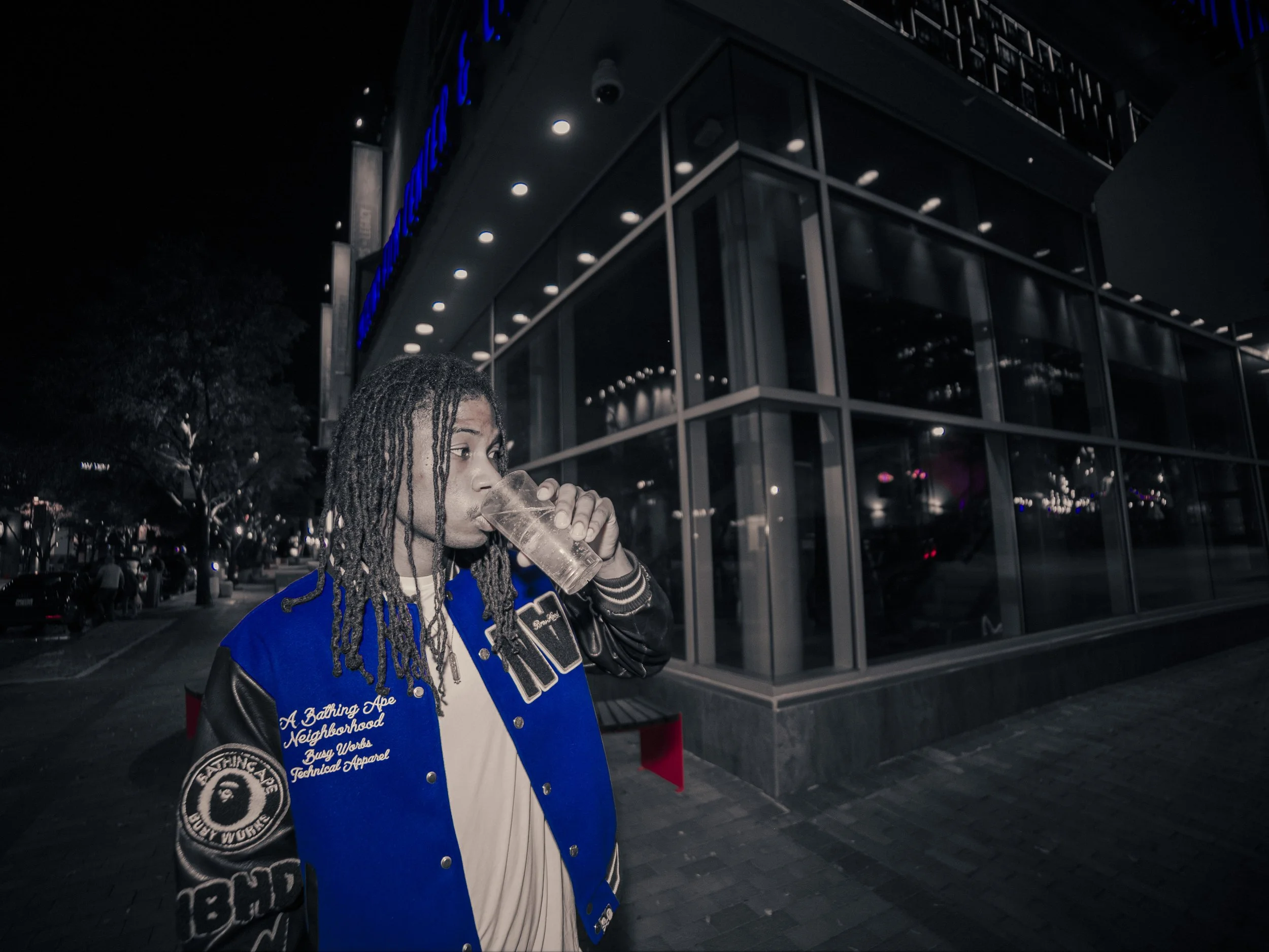 A young man with dreadlocks wearing a blue and black jacket, standing outside a modern glass building at night, drinking from a clear glass.