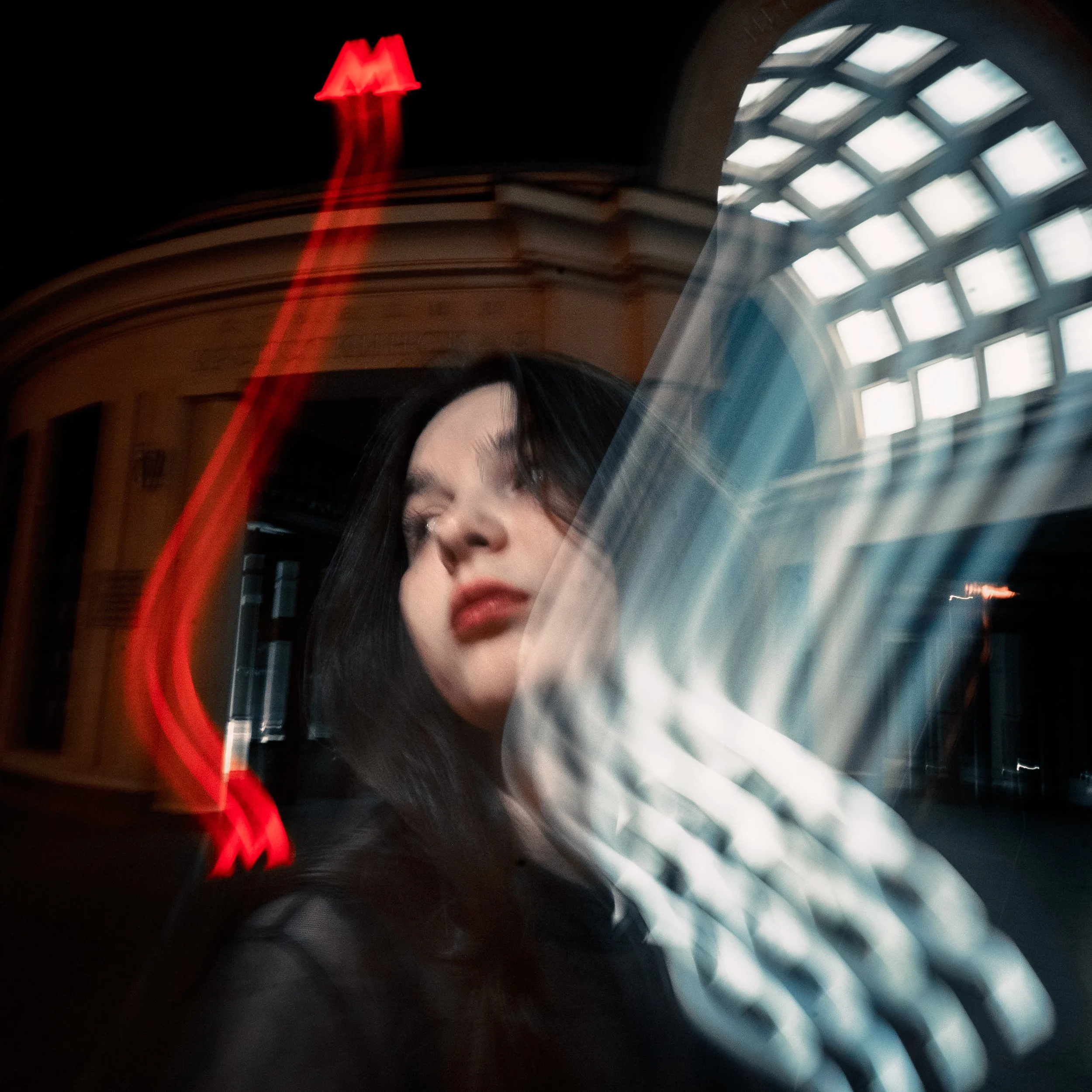 A woman with dark hair and red lipstick standing in front of a building at night with blurred light streaks, including a red neon sign.
