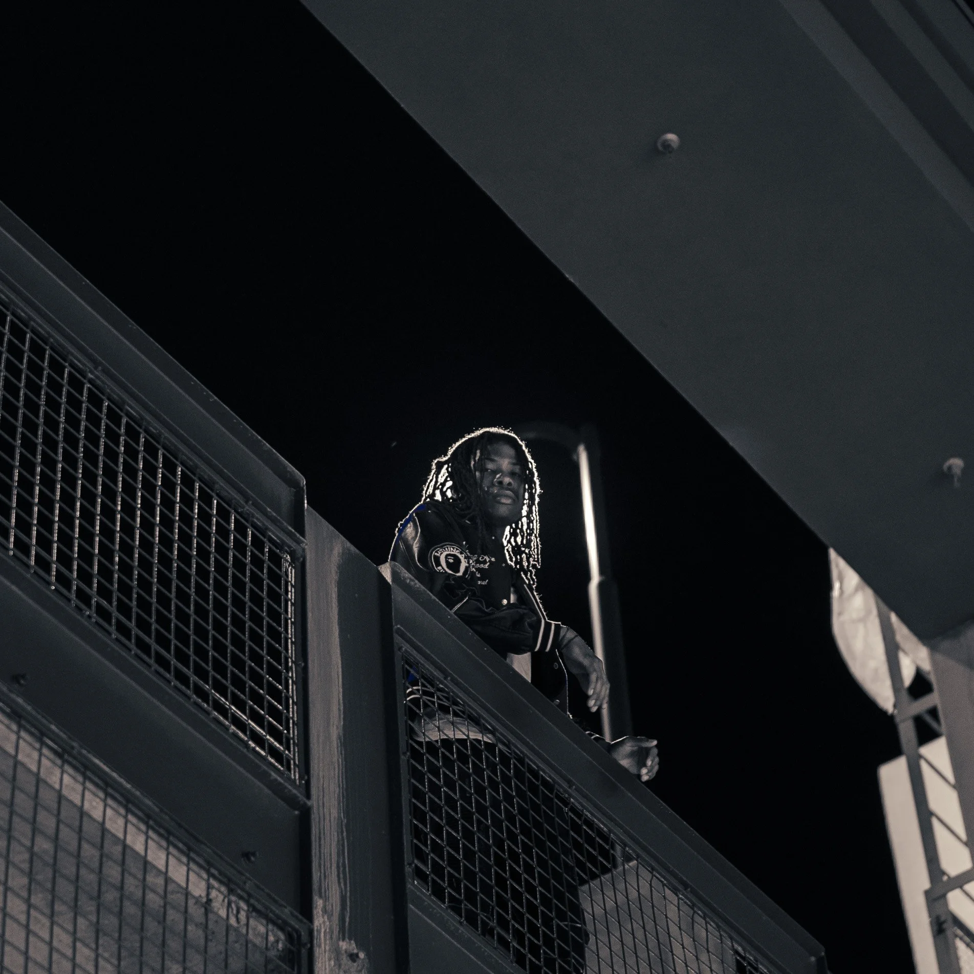 A young man with dreadlocks leaning on a railing at night, illuminated by a bright light behind him.