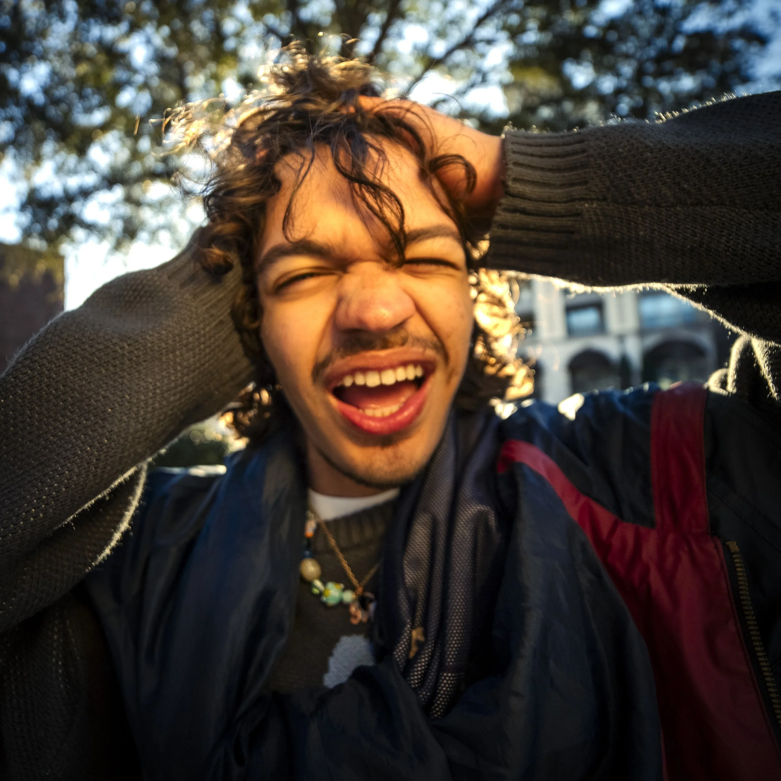 A young man with curly hair smiling and closing his eyes, holding his head with both hands, outdoors during sunset.