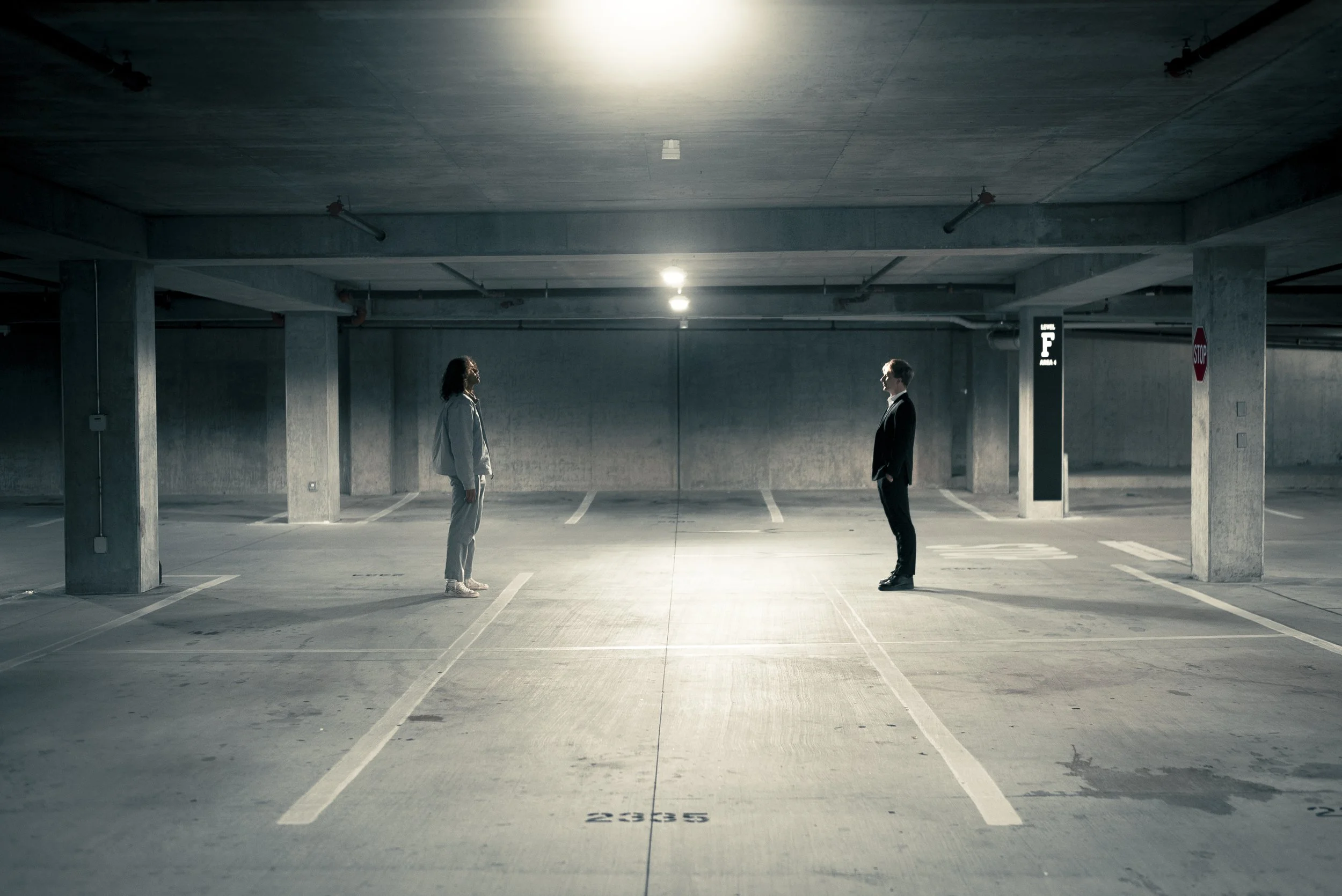 A man and woman stand facing each other in an empty, dimly lit underground parking garage.