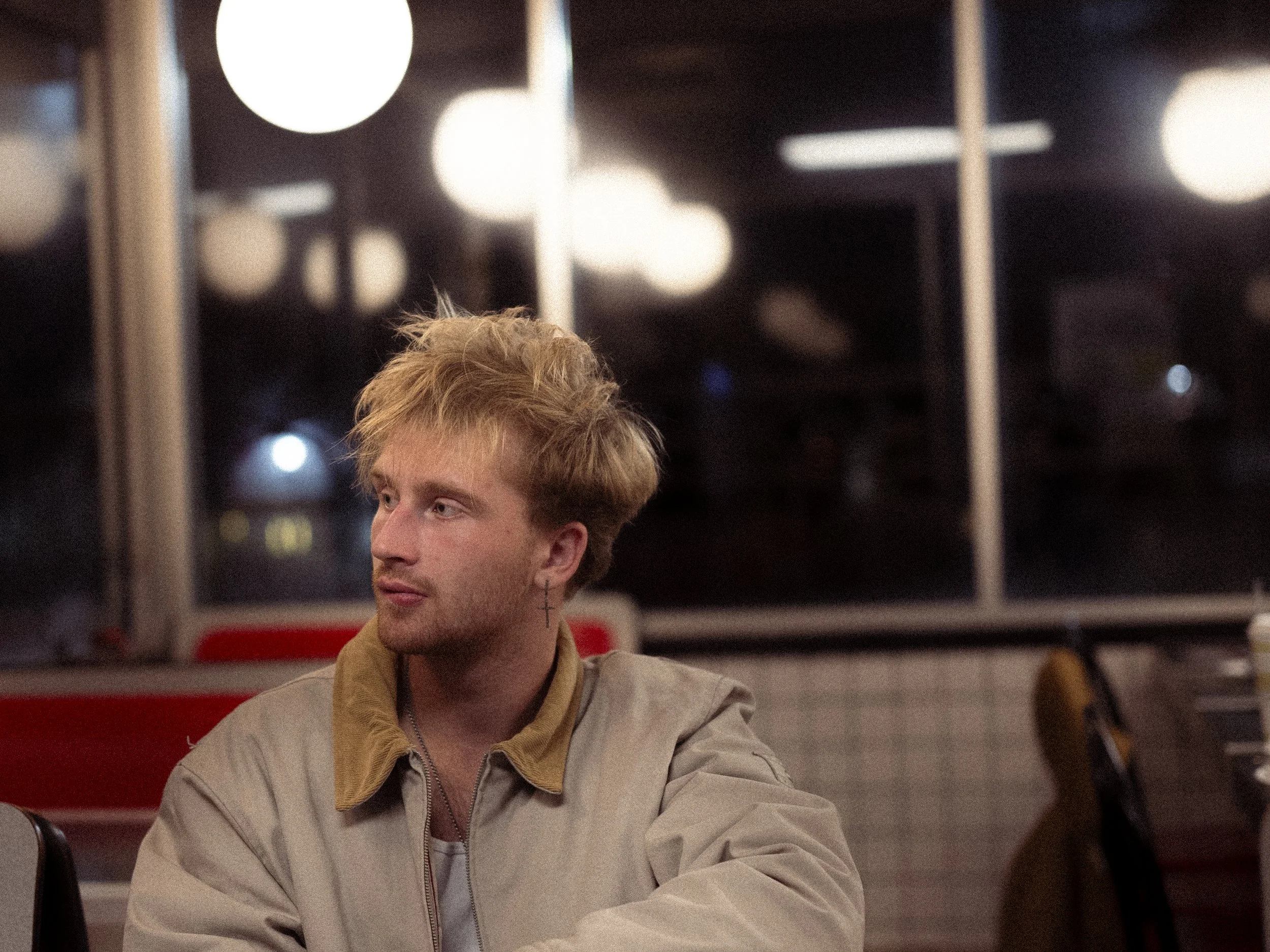 A young man with blond hair and light skin is sitting in a restaurant or diner, looking to the side. He is wearing a beige jacket with a tan collar, and a small cross earring is visible on his left ear. The background features large window panels with bright round lights and some blurry reflections.