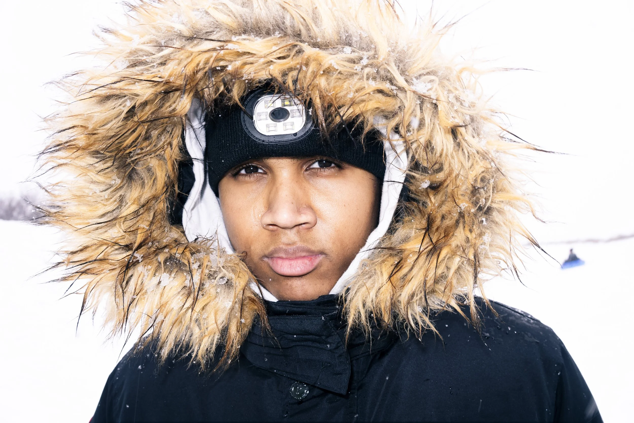 Close-up of a young man in winter clothing with a fur-lined hood, wearing a black beanie with a headlamp, against a snowy background.