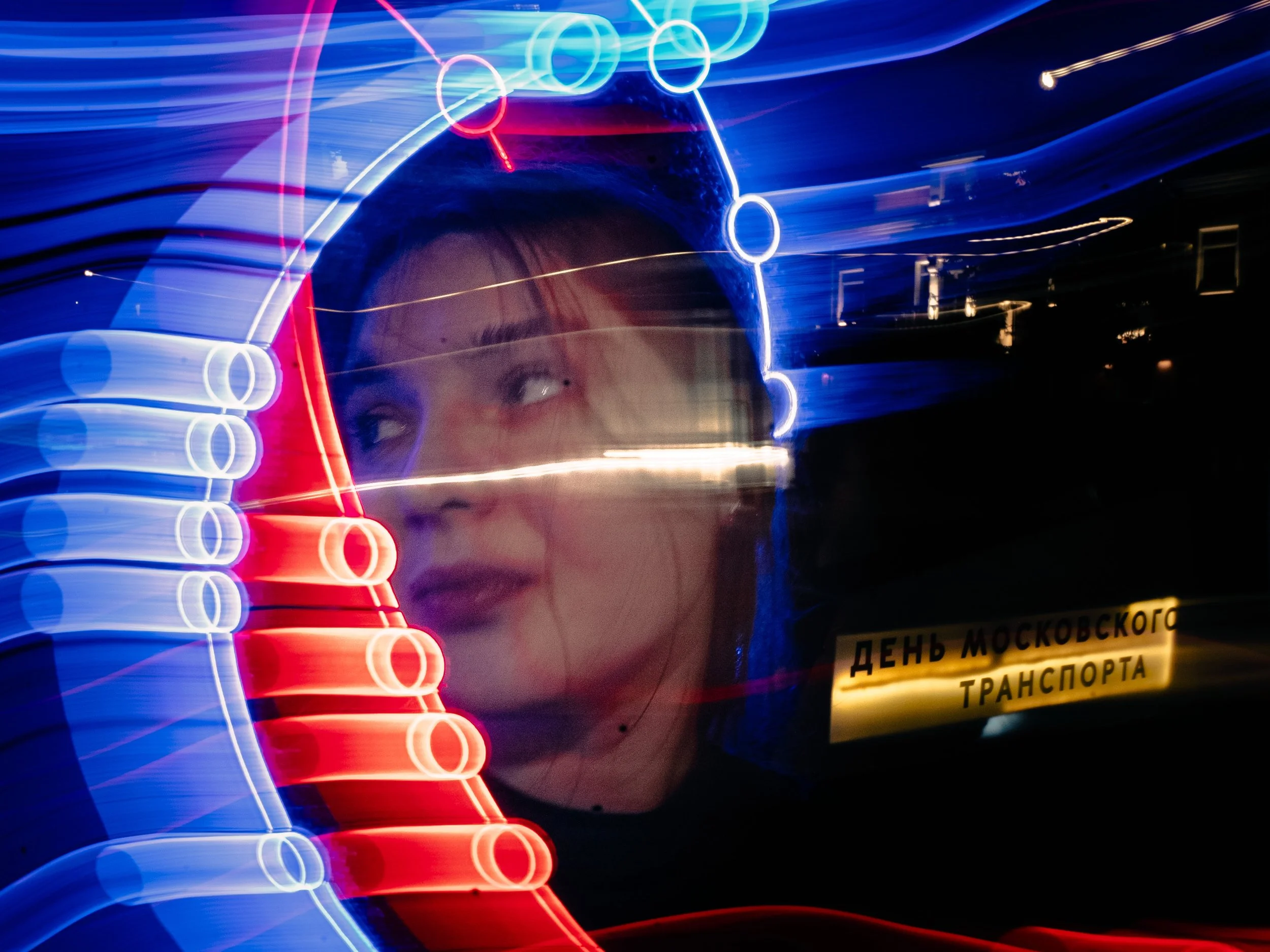 A person with a face mask looking through a neon-lit window or glass, with colorful light streaks and circular patterns, and a sign in Russian that reads "ДЕНЬ МОСКОВСКОГО ТРАНСПОРТА" (Moscow Transport Day).