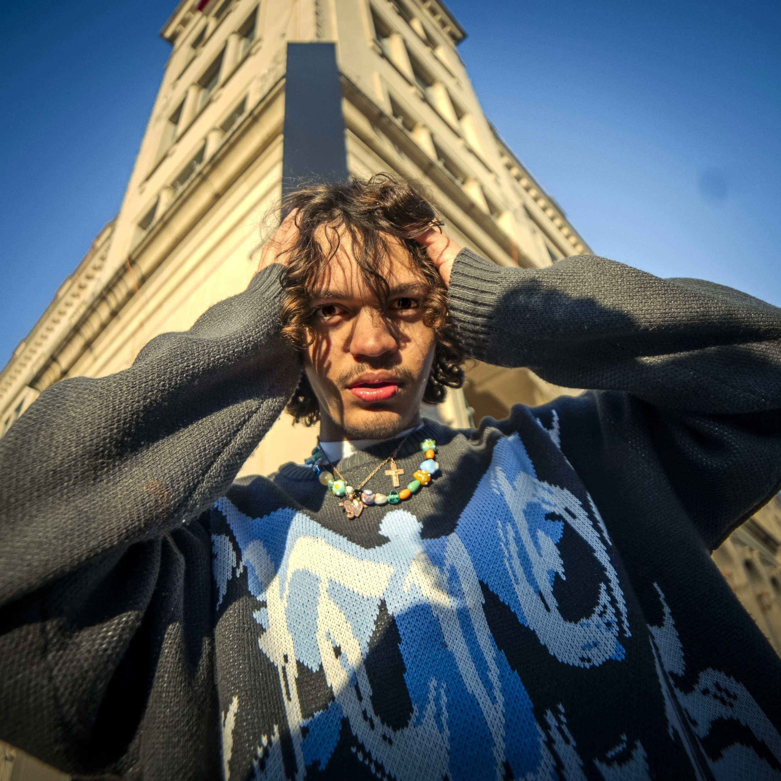 Young man with curly hair and necklaces, standing below a tall building with columns, looking directly at the camera with hands on his head during daytime.