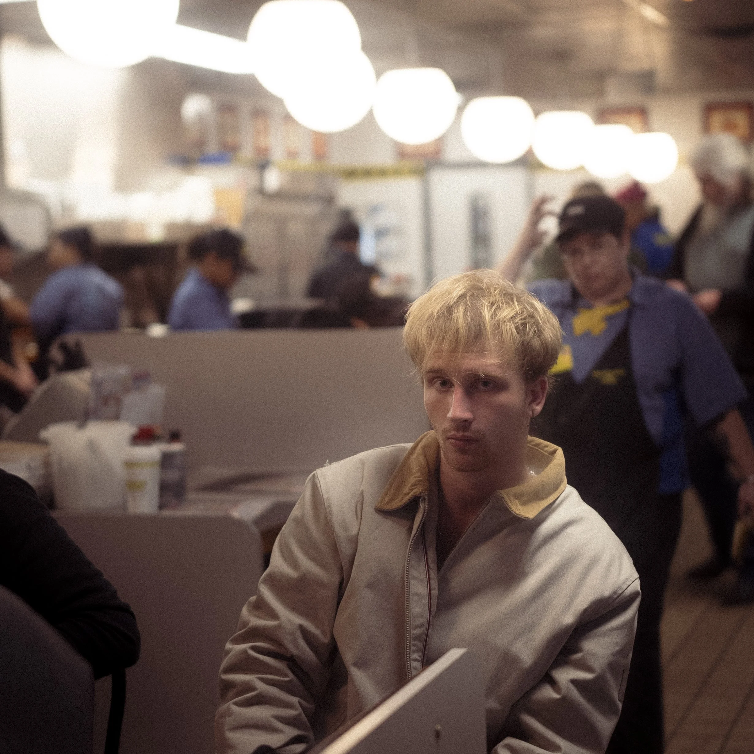 A young man with blond hair and a beige jacket sitting at a restaurant table, with a concerned or serious expression. The background shows other patrons and restaurant staff, with warm lighting and round ceiling lights.