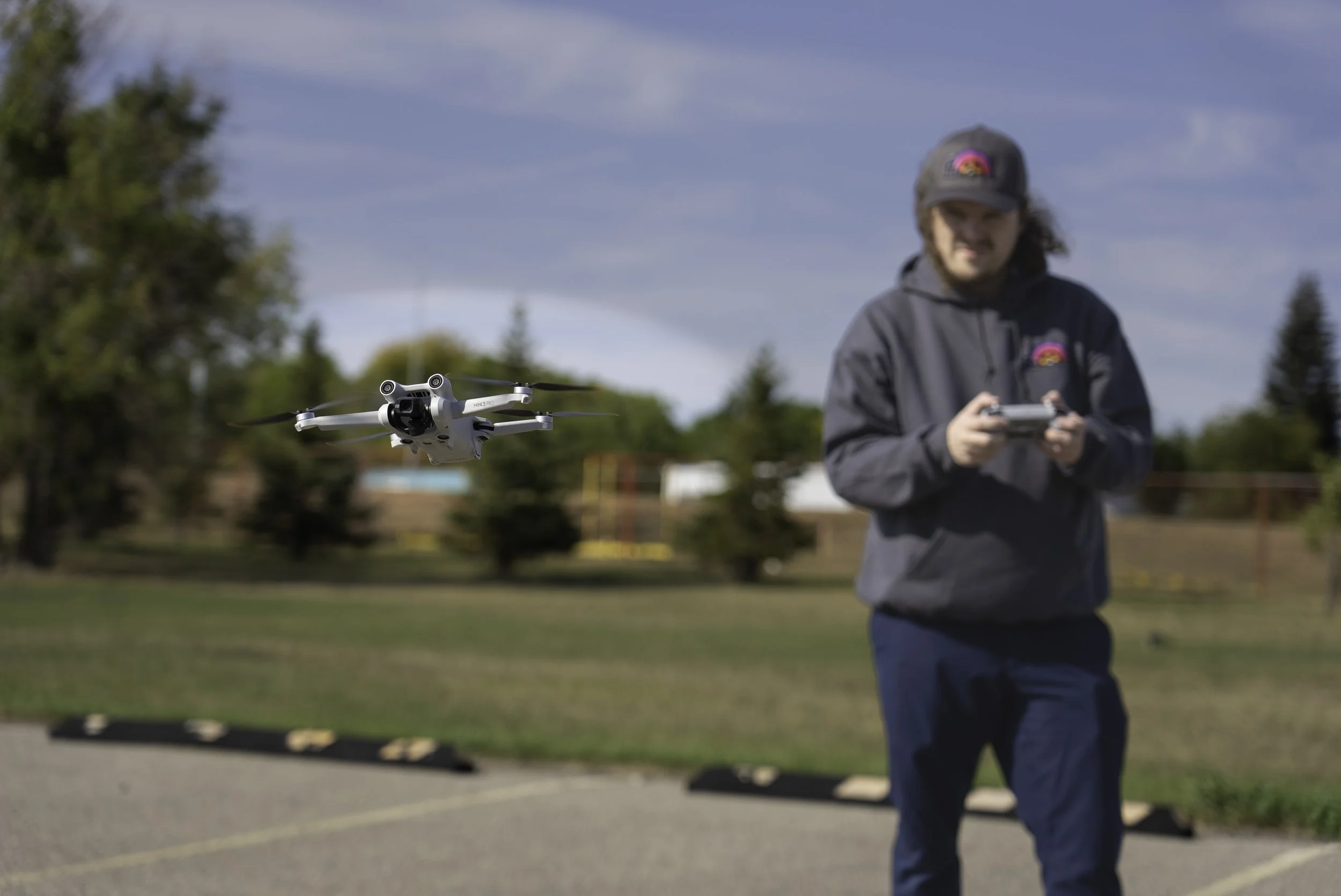 A person in a gray jacket and gray cap flying a white drone outdoors on a sunny day, with trees and a parking lot in the background.