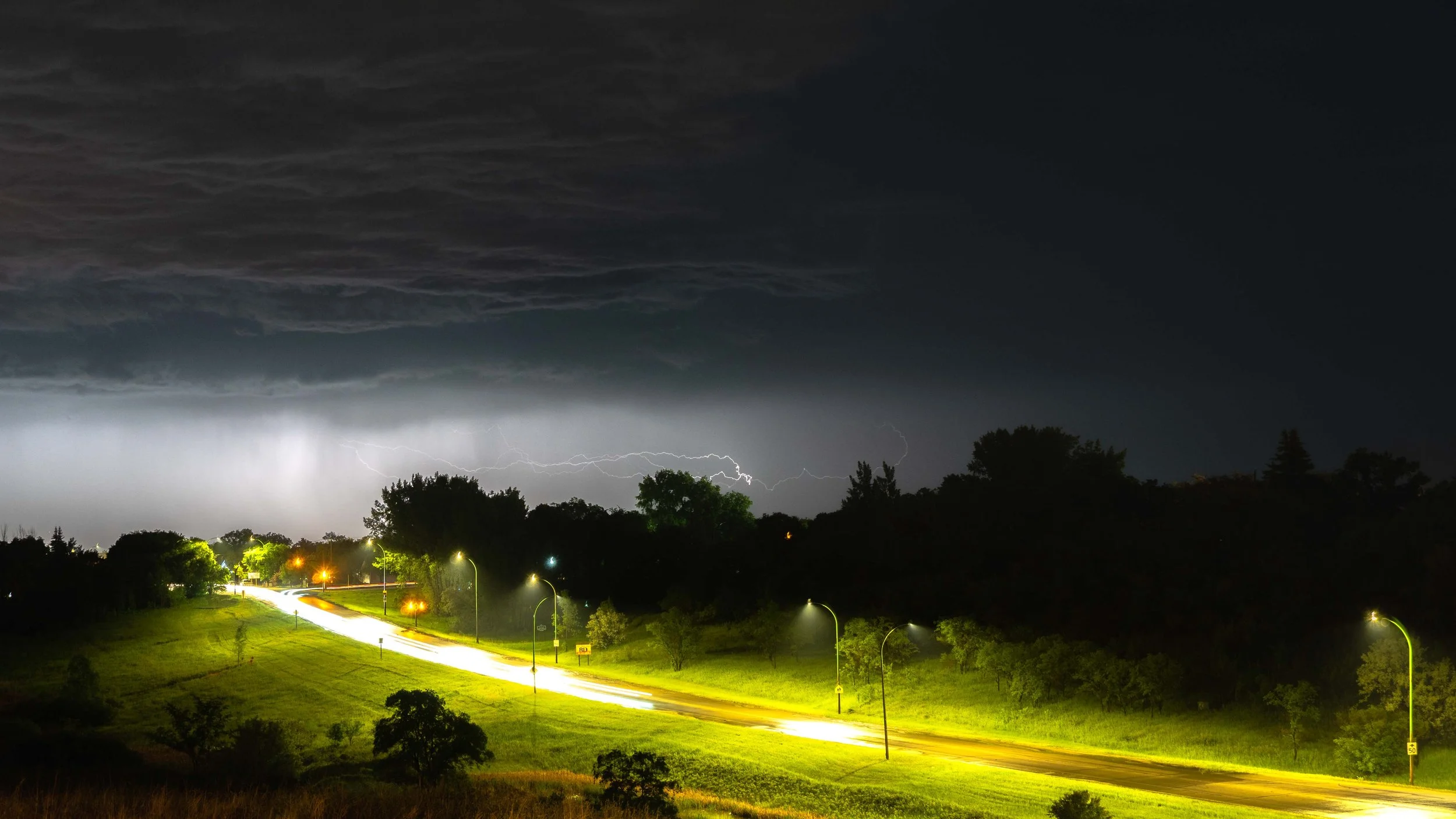 Nighttime scene of a road with streetlights illuminating the area, dark trees in the background, and lightning in the stormy sky overhead.