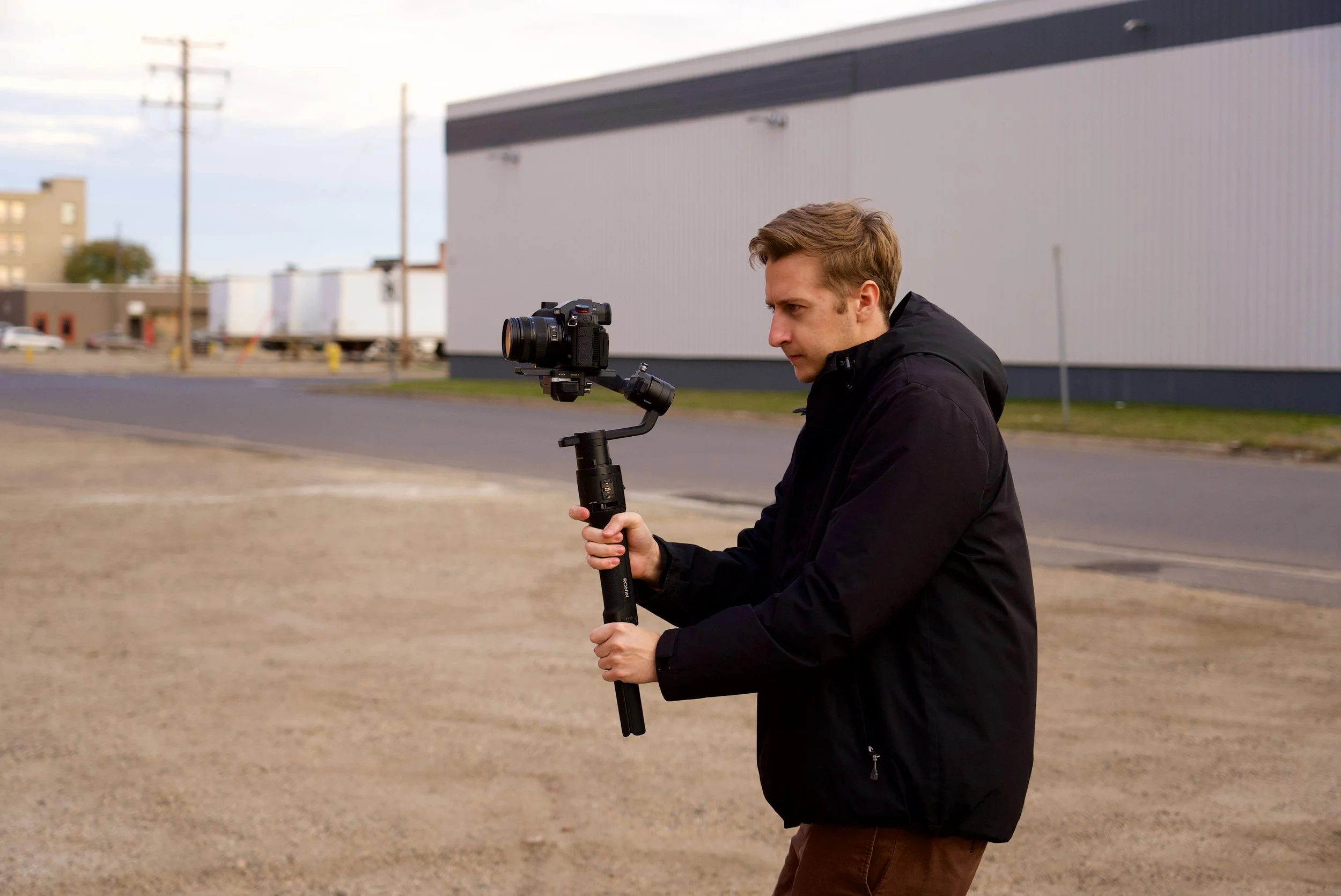 A young man in a black jacket holding a camera mounted on a stabilizer in an outdoor urban setting.